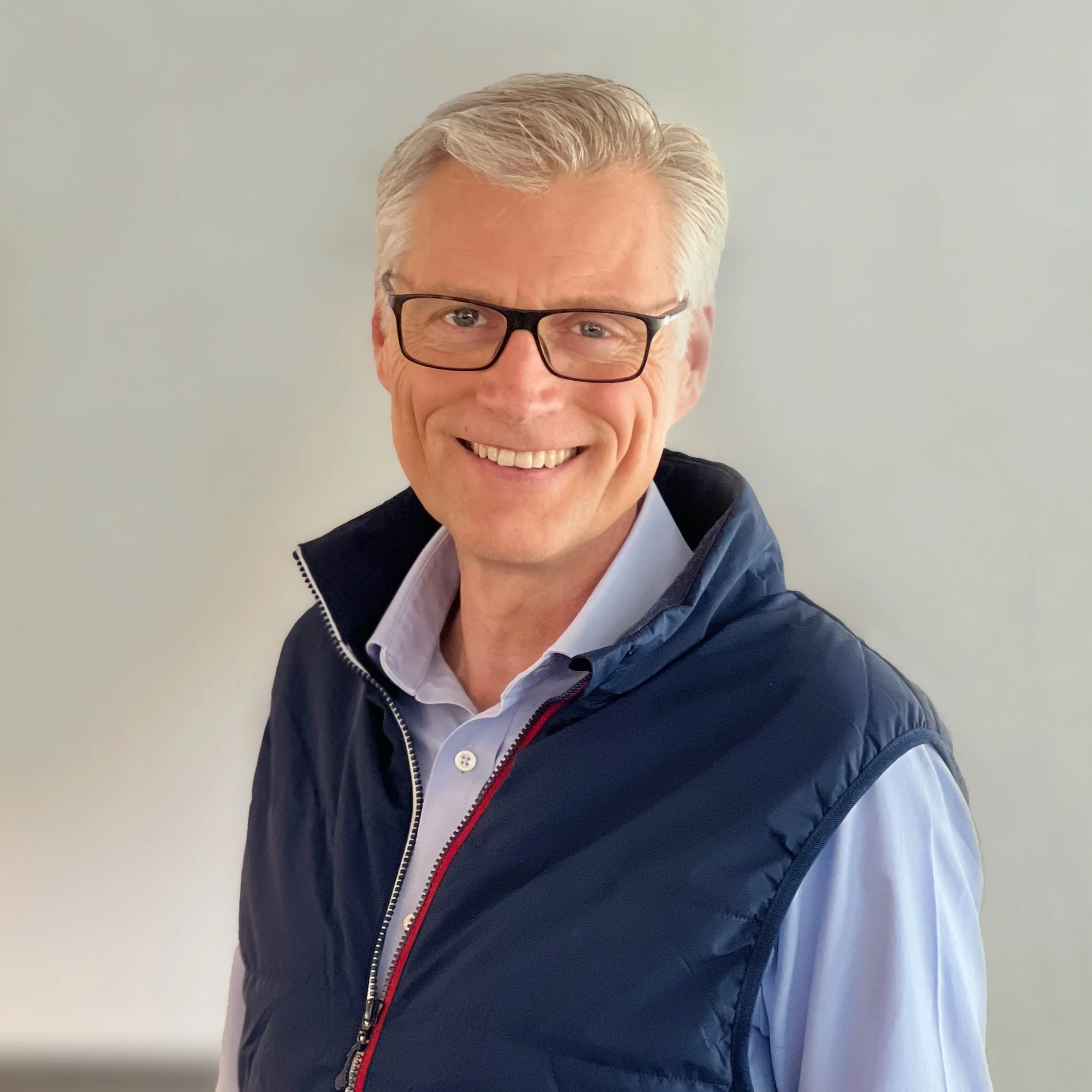 Portrait of a smiling man with gray hair and glasses, wearing a light blue shirt and a navy vest, standing against a light background.