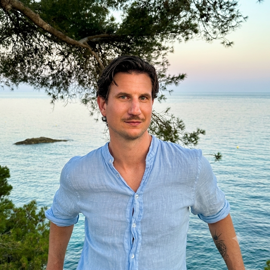 Andrew Rowe in a pale blue linen shirt, standing in front of the sea at dusk under a pine tree