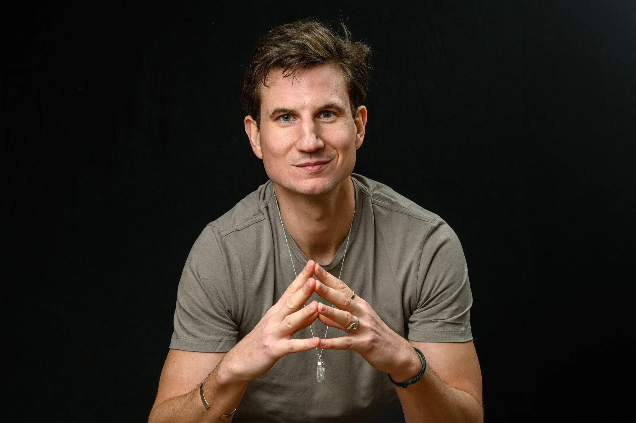 Andrew Rowe in a grey t-shirt against a black background, hands together, looking at the camera