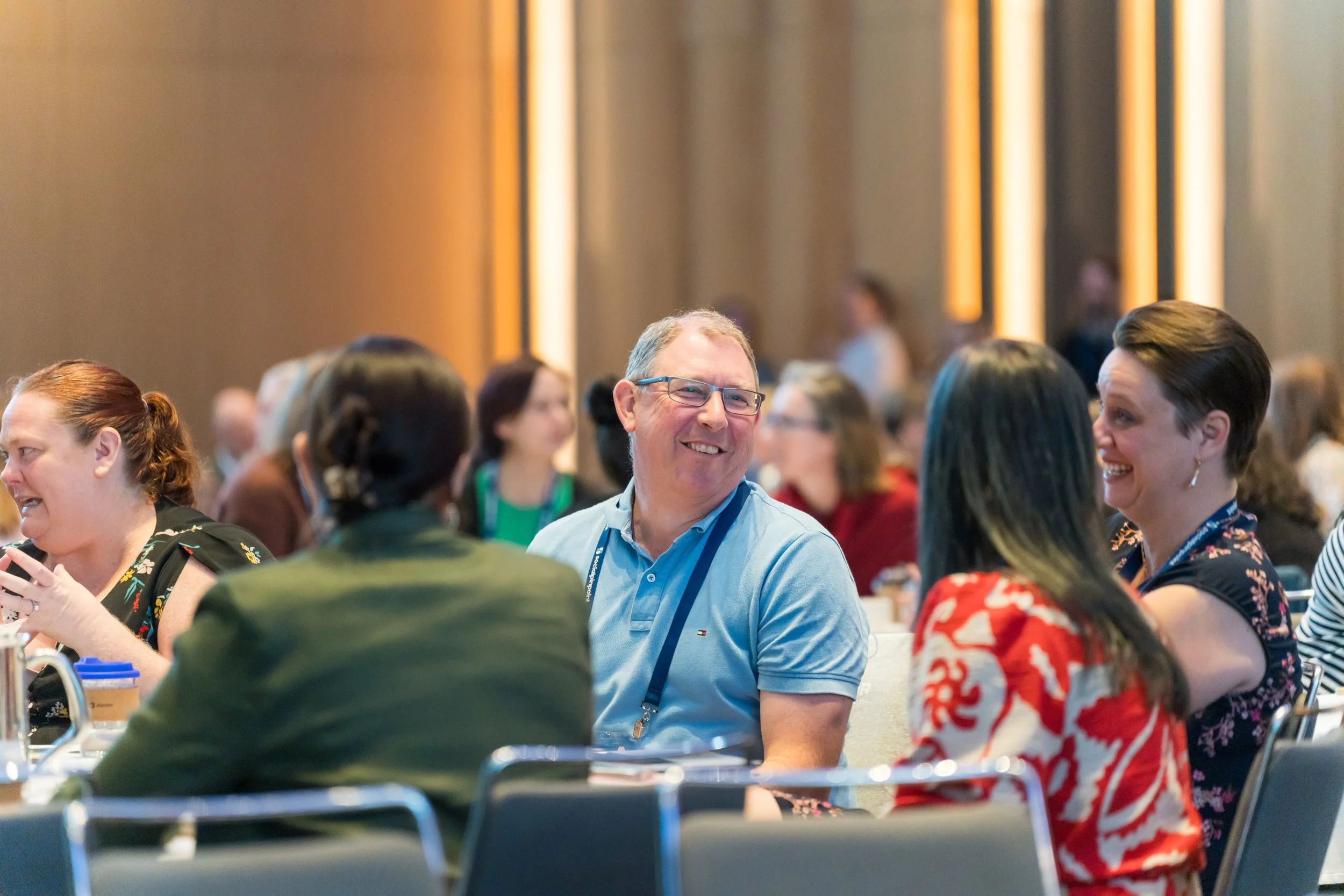 People seated at tables in a conference or event, engaging in conversation and smiling.