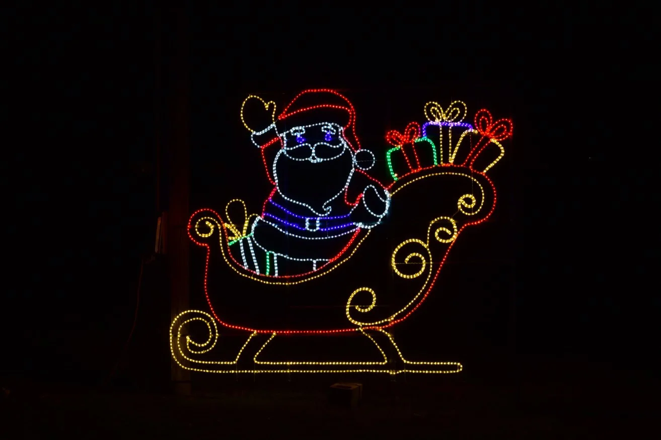 Colorful Christmas light display of Santa Claus riding in a sleigh filled with presents, outlined with bright lights against a dark background.