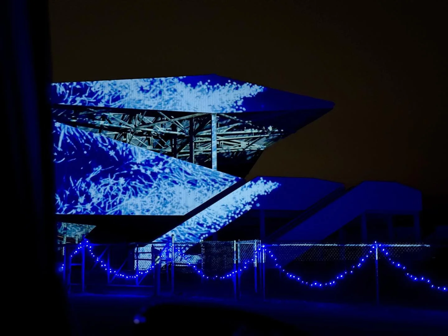 Night view of a modern building with illuminated blue digital display on its exterior, featuring abstract patterns and structural framework.