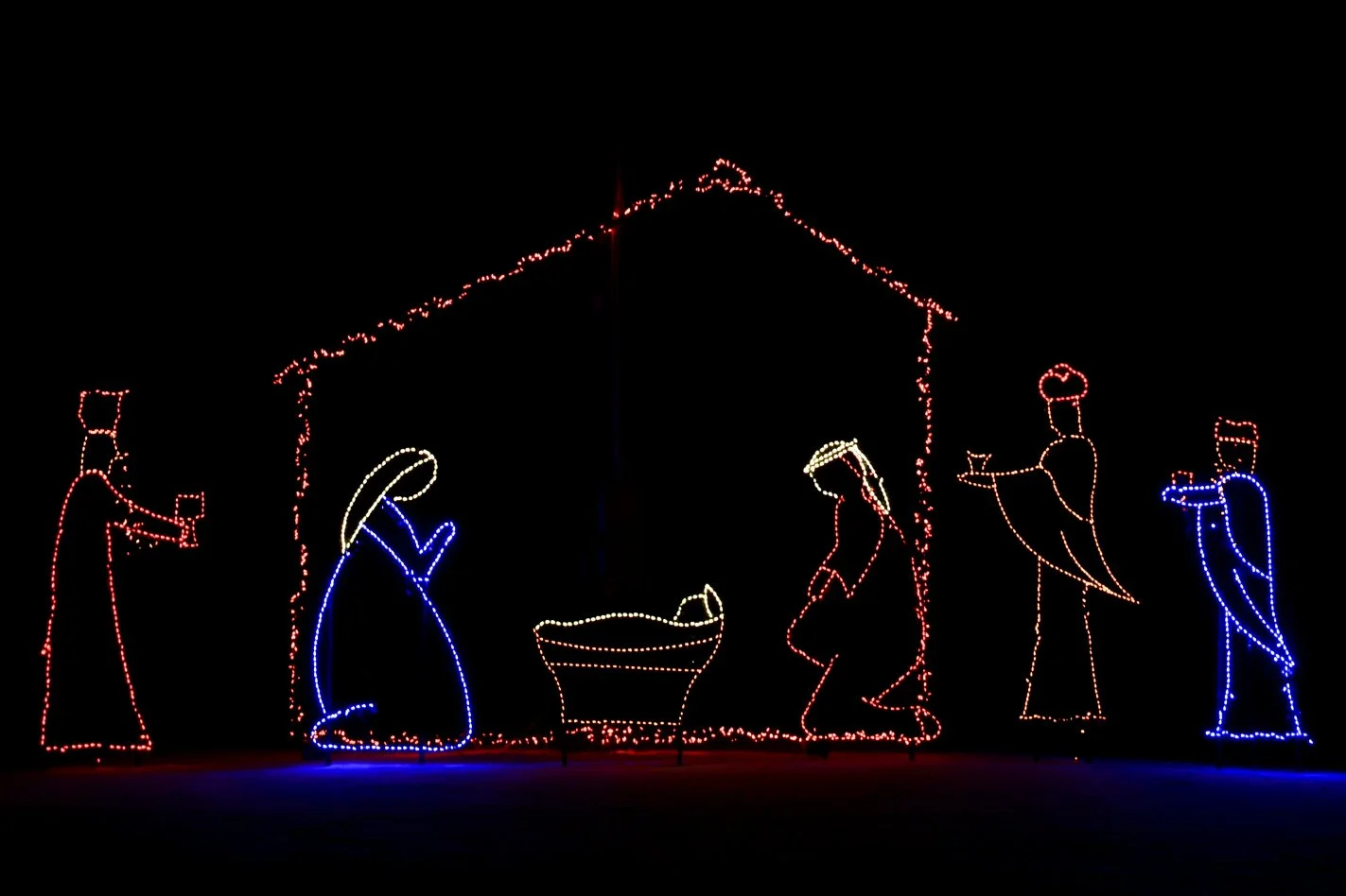 Christmas nativity scene outline made of colorful lights, with figures of Mary, Joseph, baby Jesus, angels, a shepherd, and animals, set against a dark background.