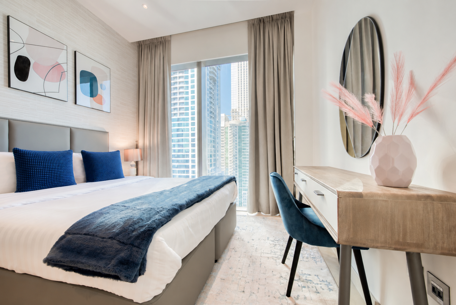 Dubai bedroom with blue cushions and throw, pastel abstract prints, a wooden desk and a pink vase beneath an oval mirror.