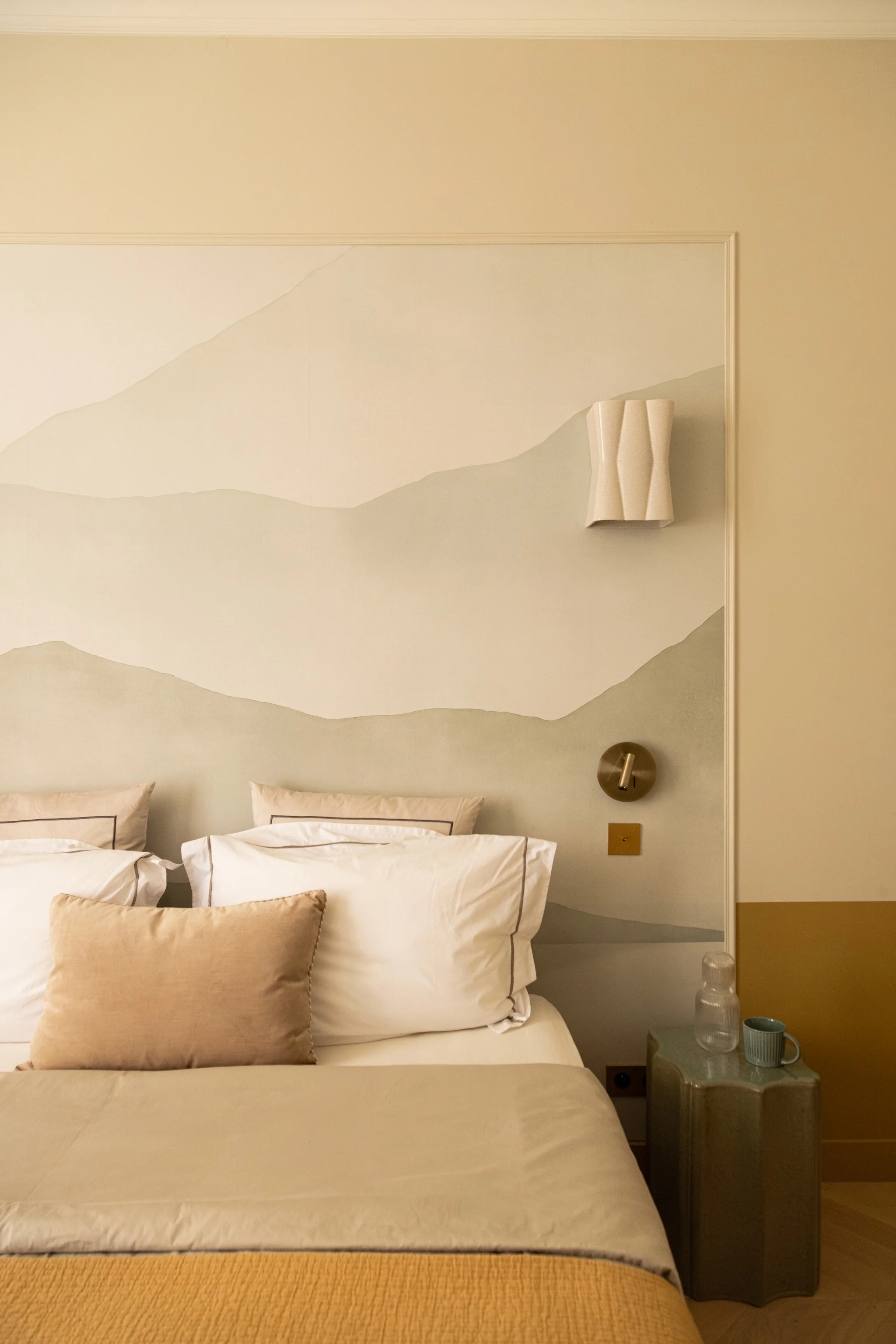 Parisian bedroom with a pale landscape wallpaper, ceramic wall sconces by Maison Sarah Lavoine, cream bedding, an ochre throw and a sculptural green bedside table.
