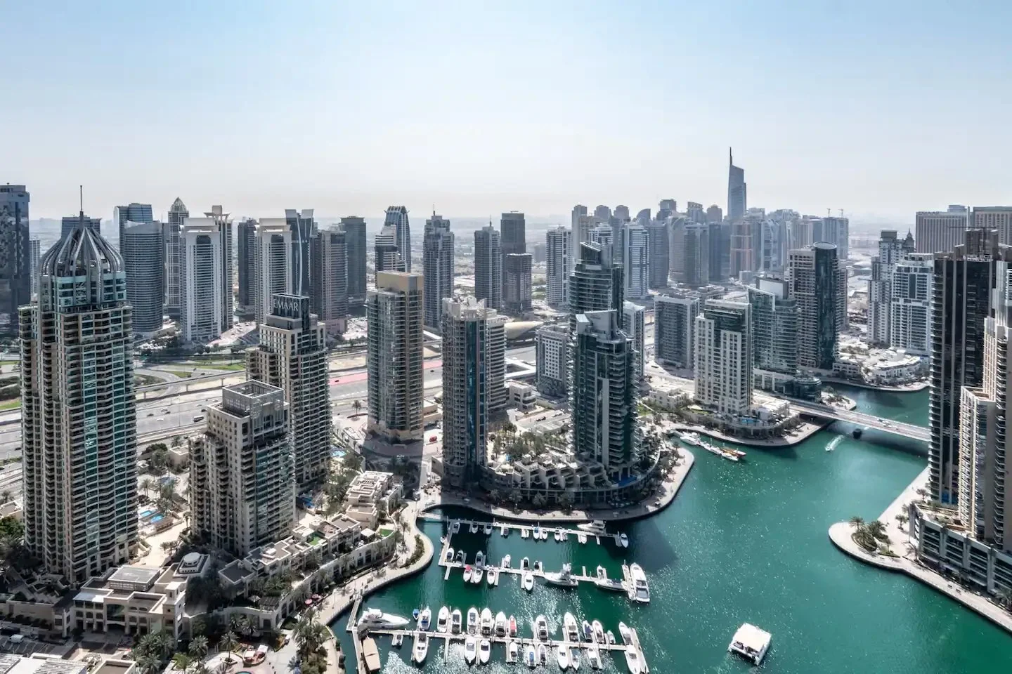 Panoramic Dubai Marina view with waterfront towers, turquoise water, moored yachts and a bridge.