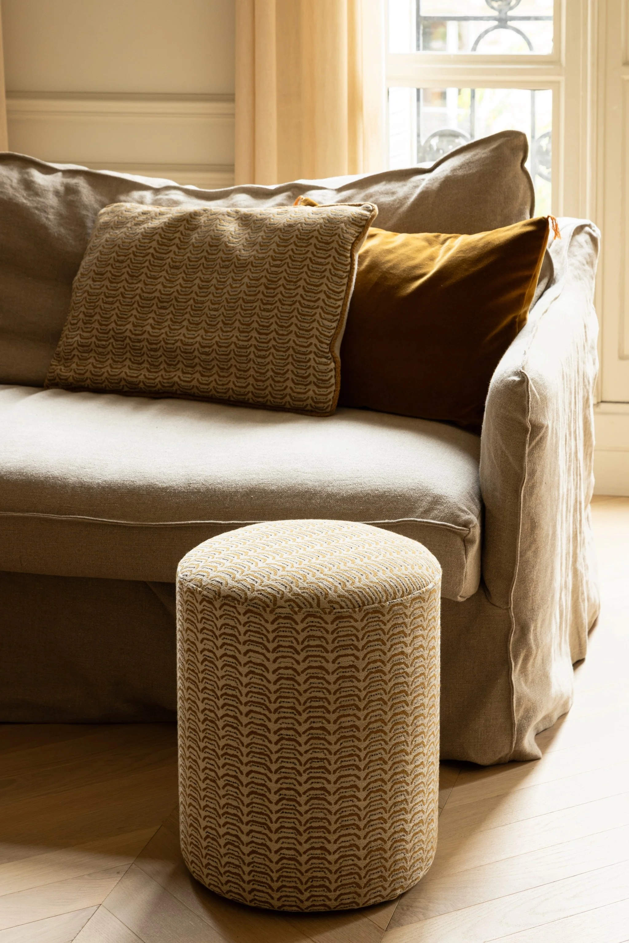 Cream linen sofa with an ochre cushion and a patterned cushion matching a cylindrical footstool by Nobilis, lit by the adjacent French window.