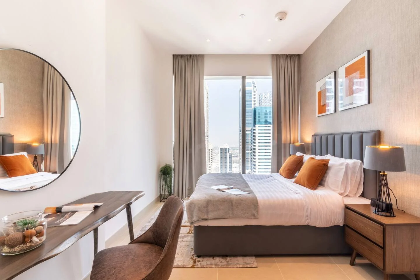 Dubai Marina bedroom with a grey panelled headboard, burnt-orange cushions, round mirror and slim wooden desk.
