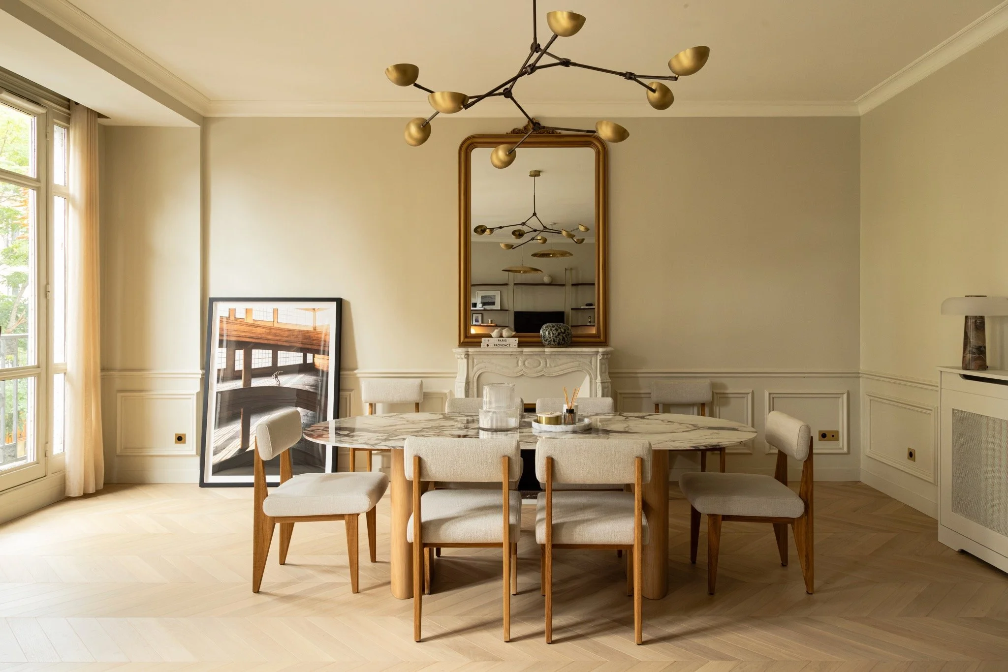 Parisian dining room with an oval veined table, cream upholstered chairs, an ornate fireplace and a branching gold-toned chandelier by 101CPH.