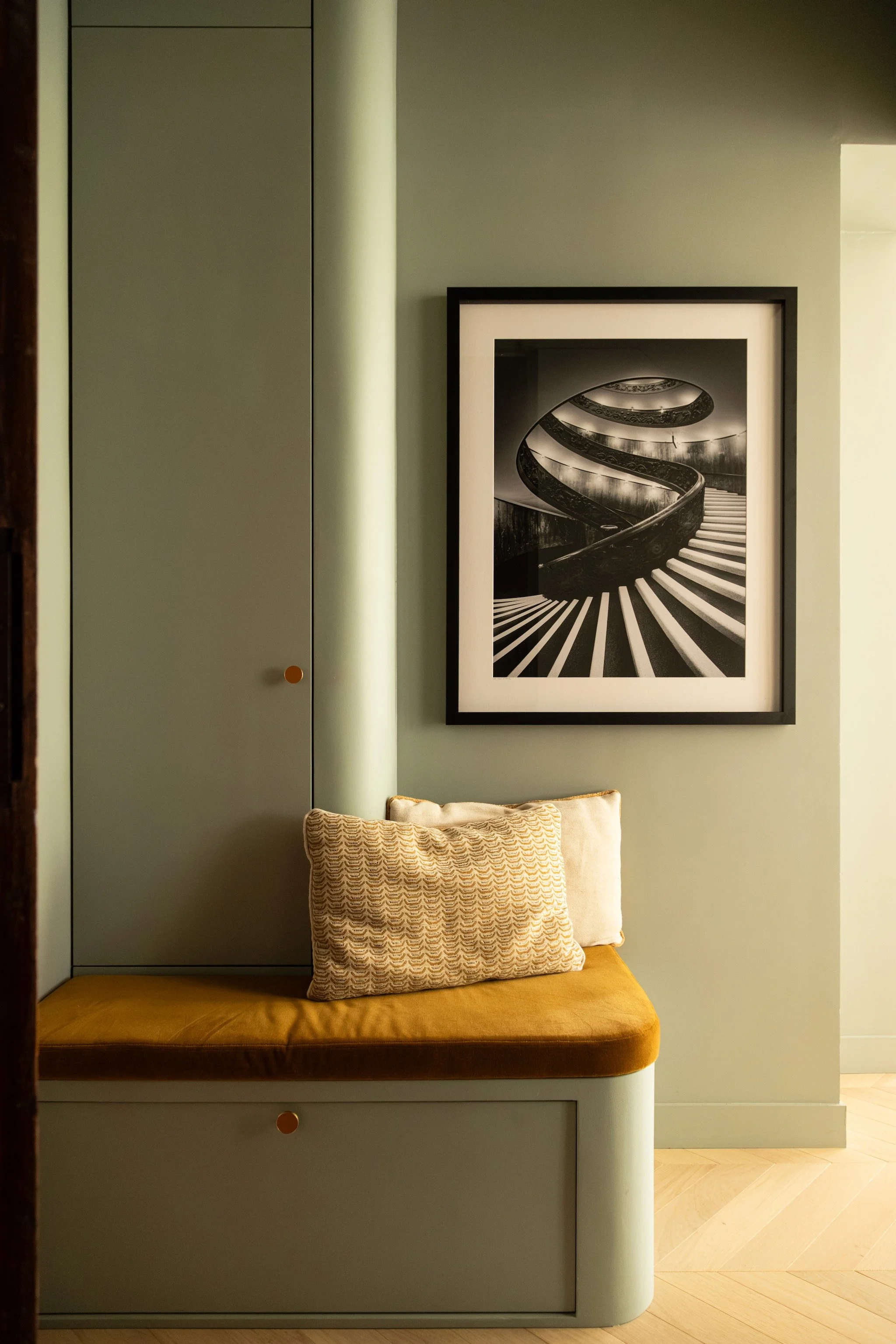Sage-green built-in storage and a curved bench with an ochre seat beneath a black-and-white staircase photograph in Paris 16th.