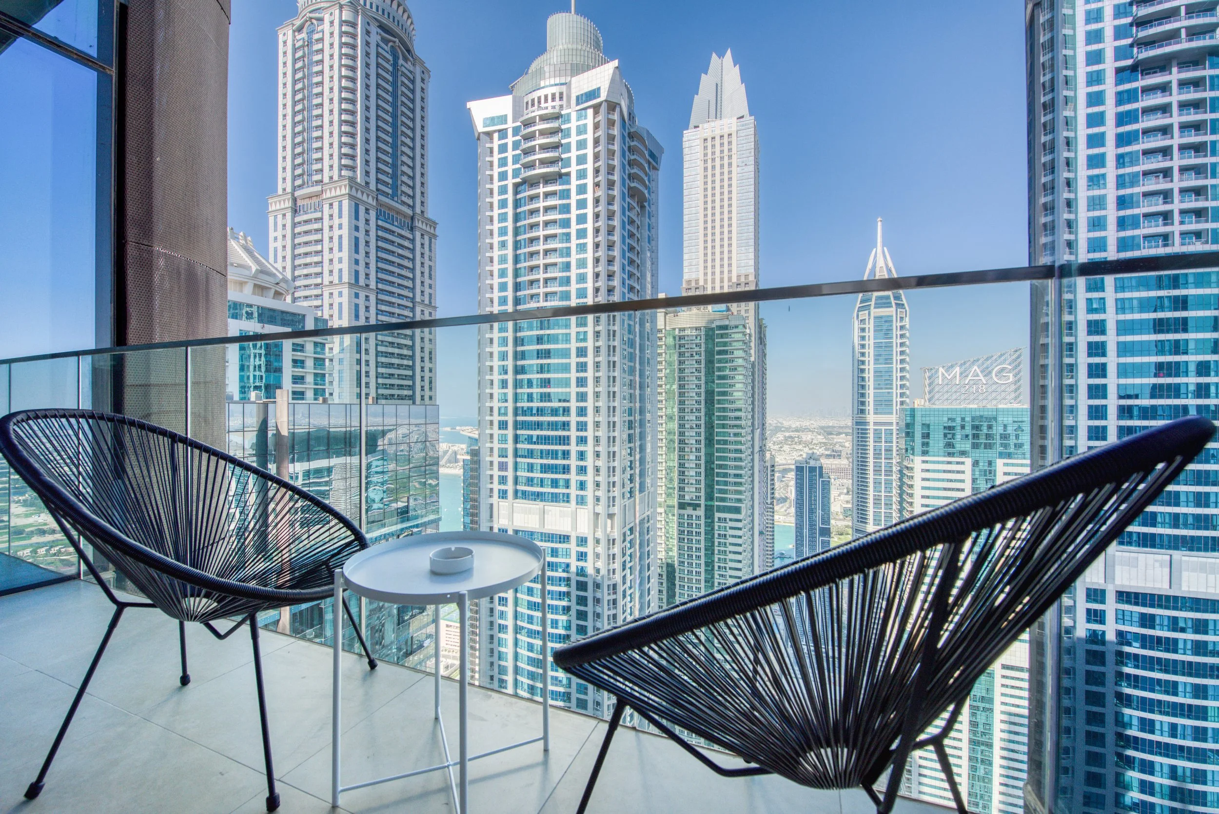 Two black cord chairs and a white round table on a glass-fronted Dubai Marina balcony overlooking neighbouring towers.