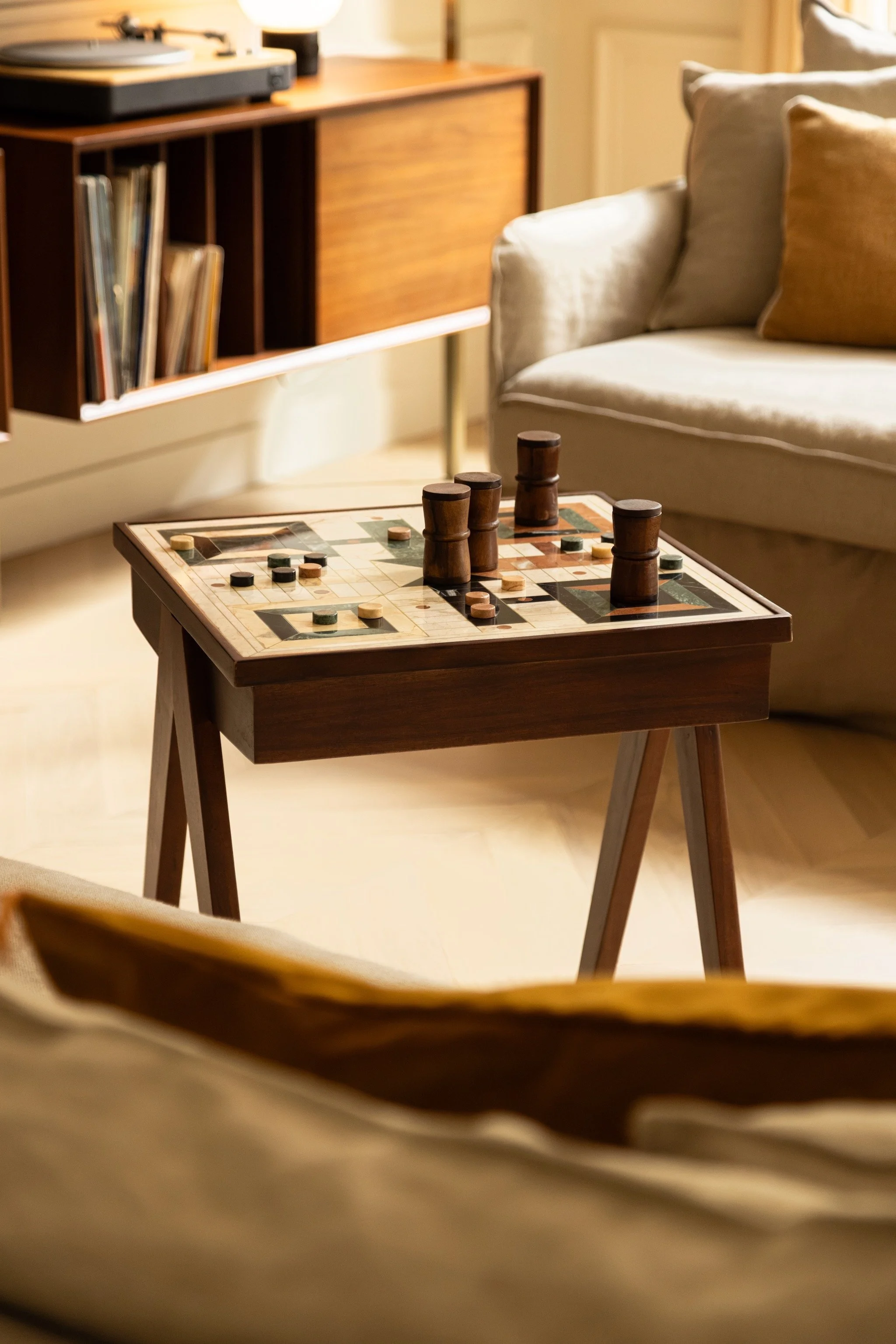 Wood and marble games table with a patterned board and playing pieces, beside a cream sofa and record cabinet in a Parisian apartment.