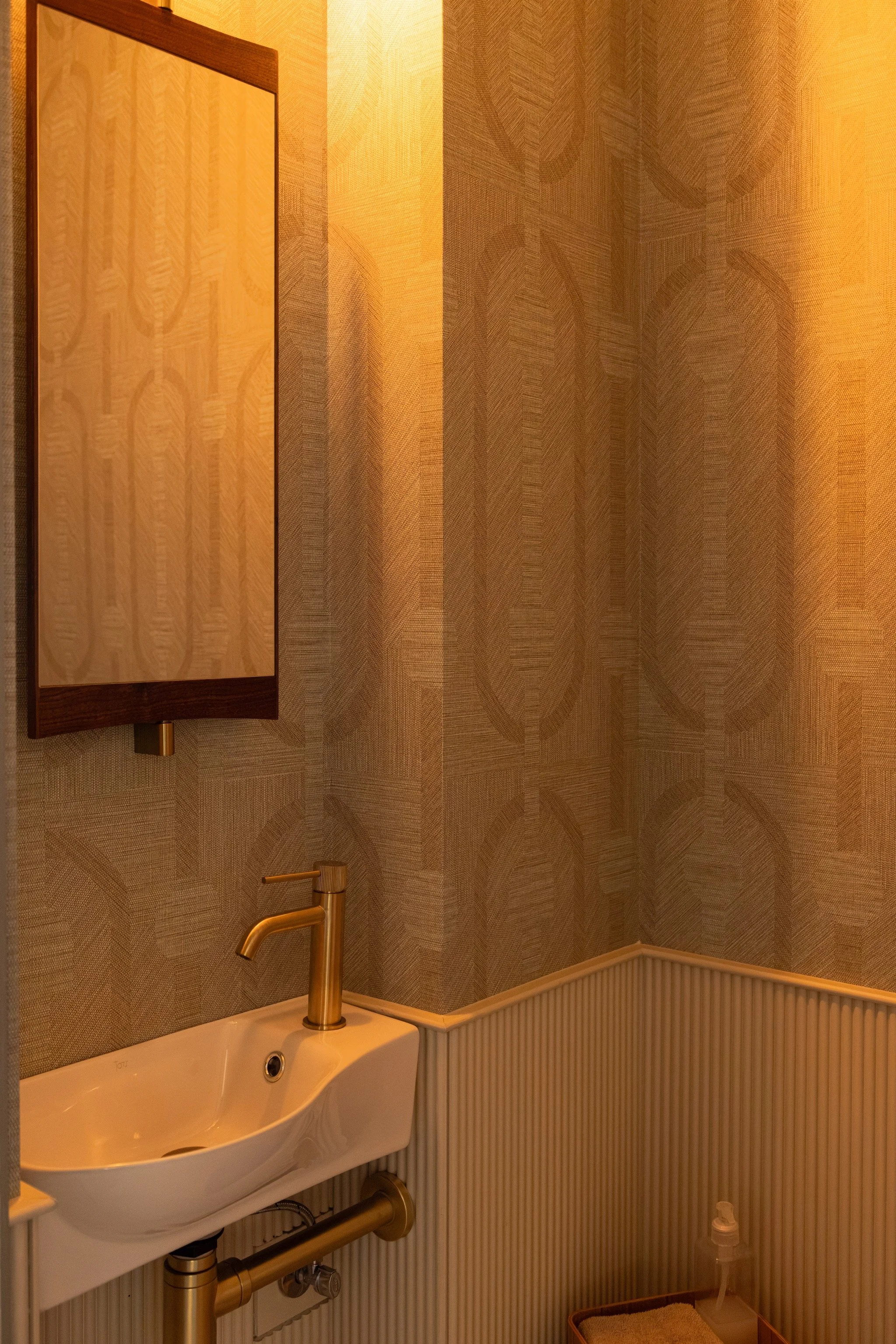 Compact washbasin with a gold-toned tap, wood-framed mirror by Gubi, leaf-patterned wallcovering by Arte and vertically ribbed lower walls by Orac.
