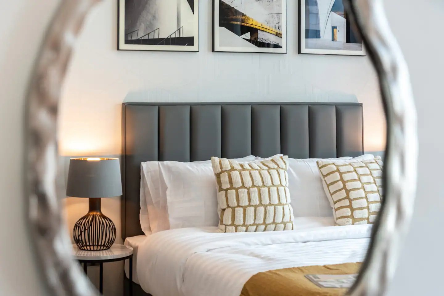 Bedroom reflected in an irregular mirror, with a grey panelled headboard, textured cream cushions and architectural prints.