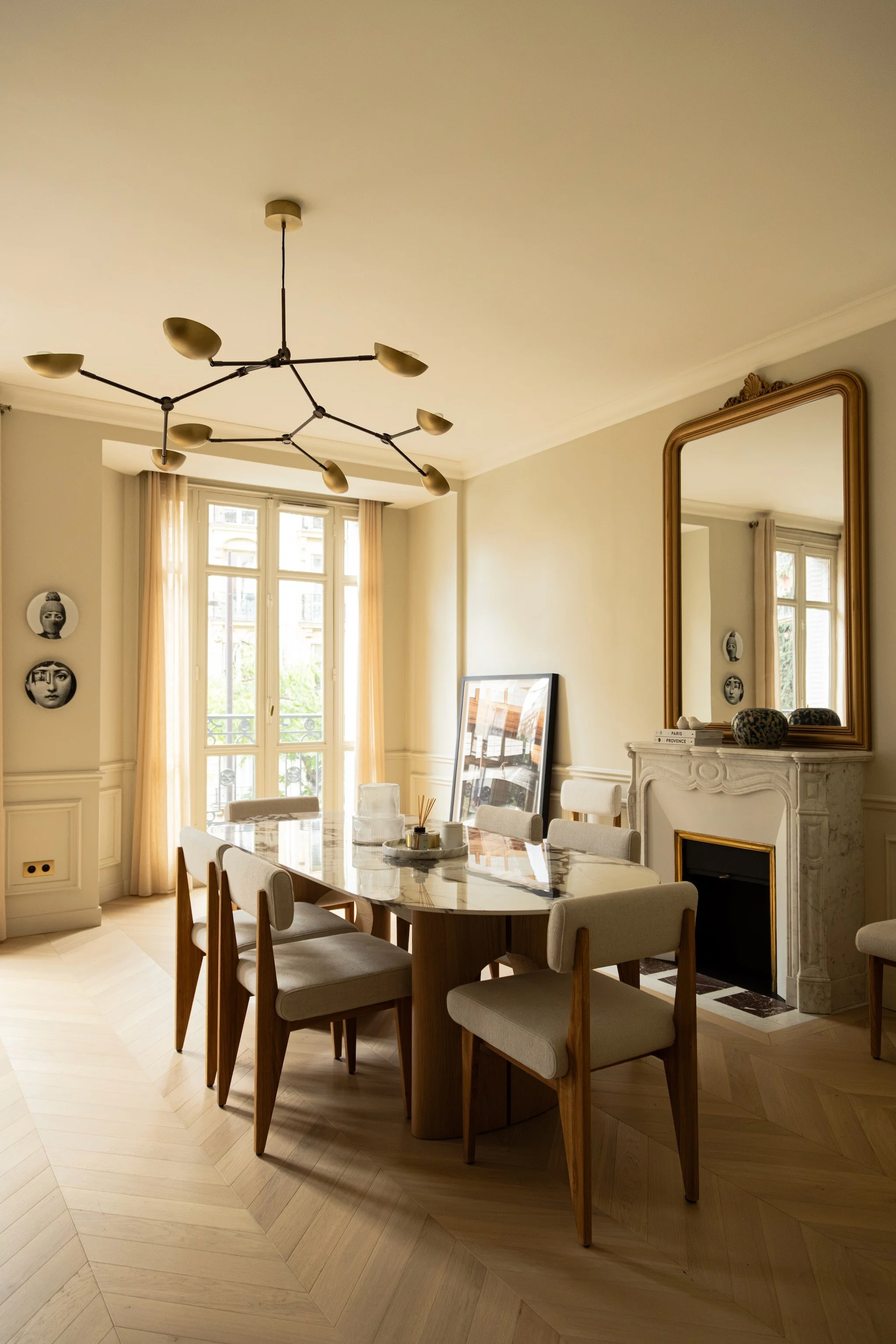 Dining area in Paris 16th with a veined oval table by AMPM, wooden chairs, tall French windows and a gold-framed mirror above the fireplace.