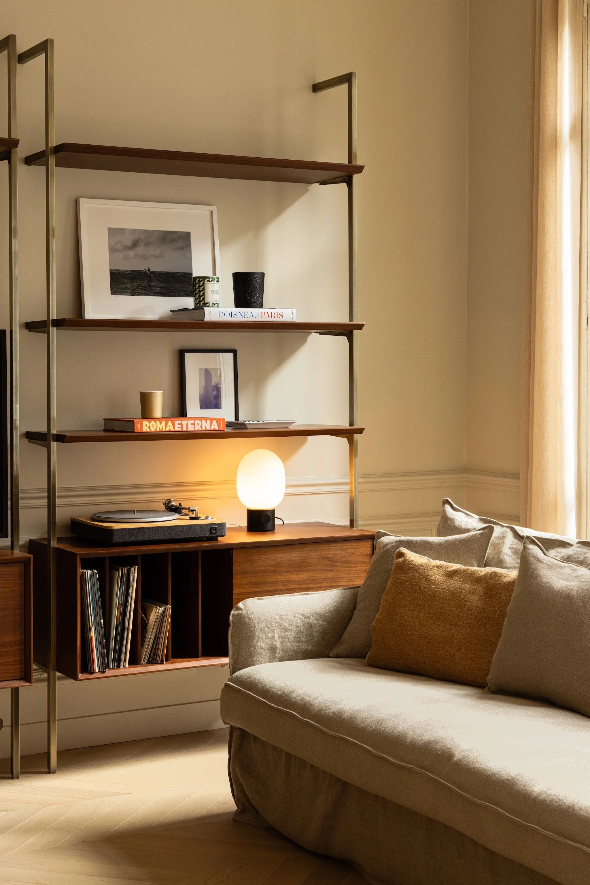 Sunlit sofa beside wooden shelving with a record player, vinyl storage, framed photographs and a glowing oval lamp, in Paris 16th.