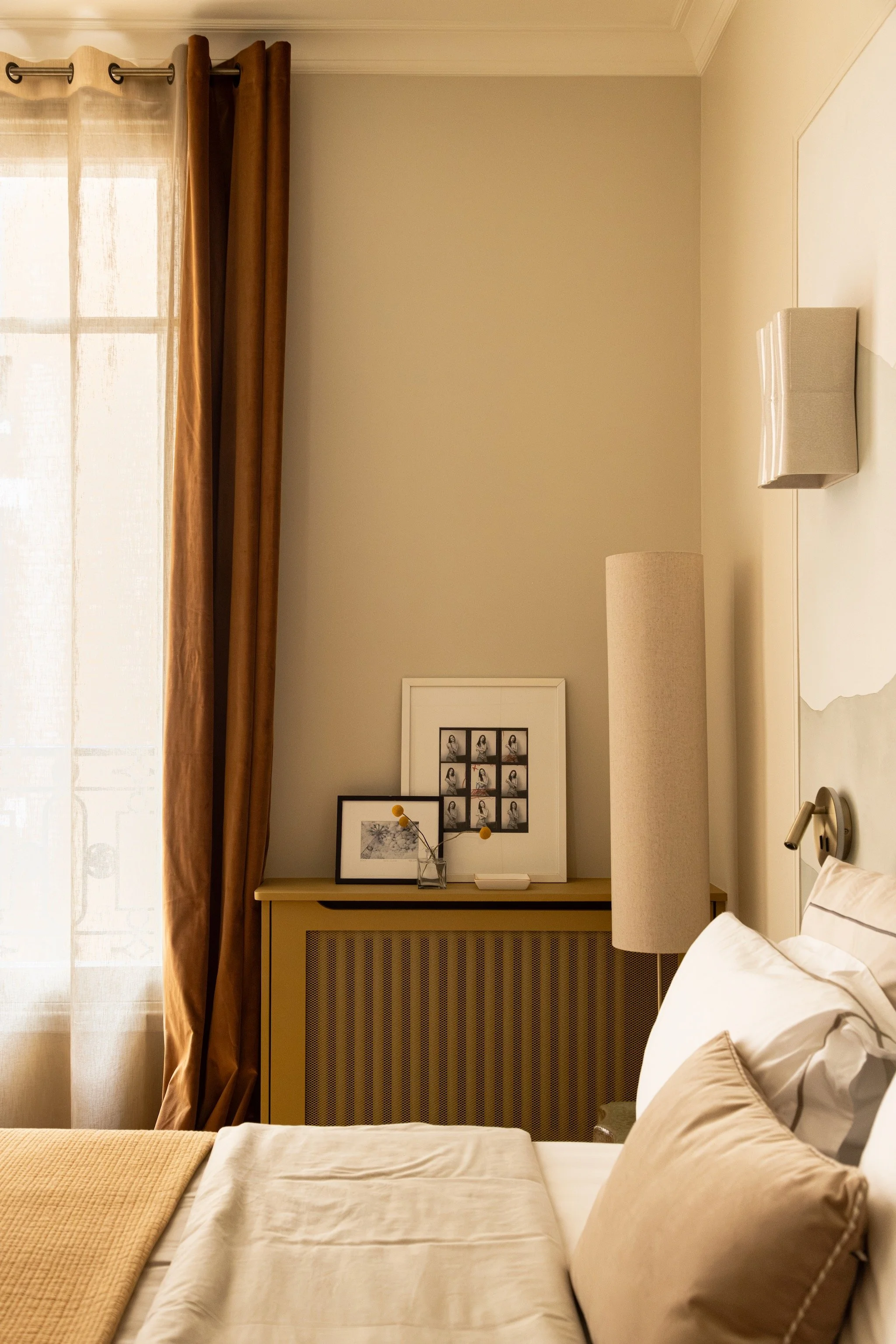 Bedroom corner with rust-coloured curtains, an ochre radiator cover, framed photographs and a tall cylindrical floor lamp.