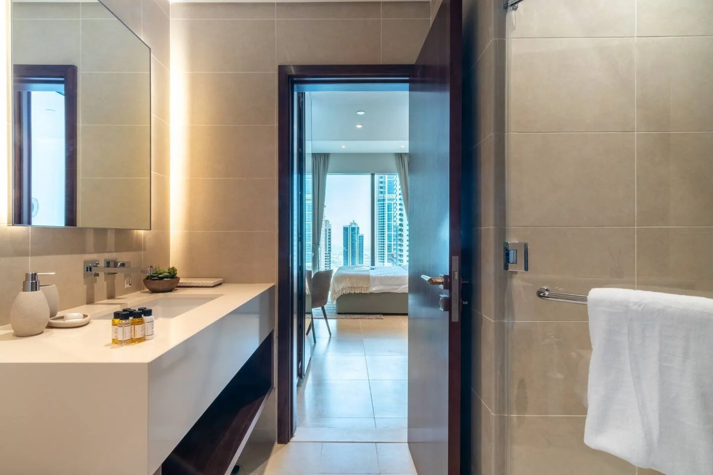 Beige-tiled bathroom with a white vanity, illuminated mirror and glass shower screen, opening onto a bright bedroom.