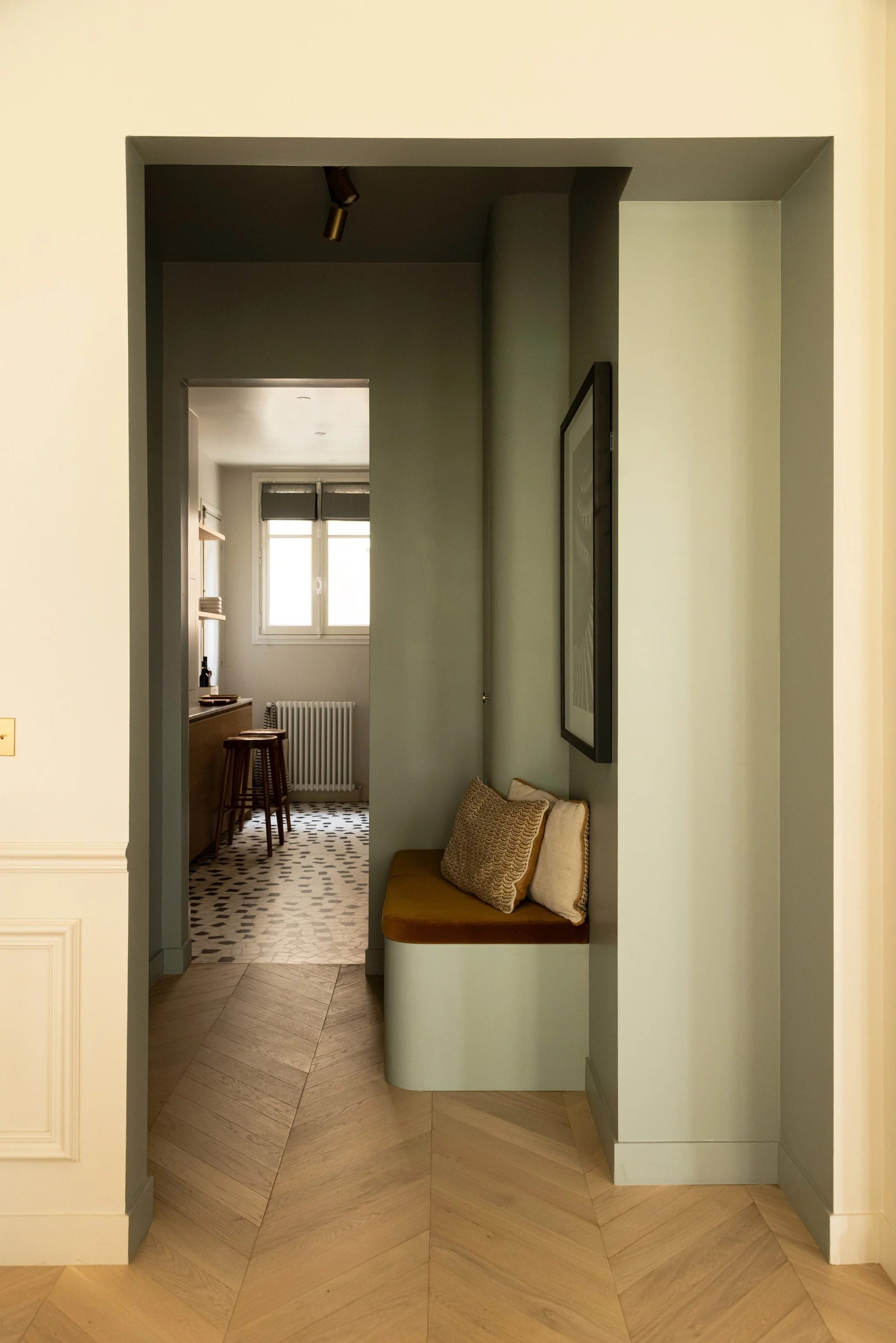 Sage green entrance hall with built-in joinery and an ochre velvet cushion by Pierre Frey, showing the kitchen in the background.
