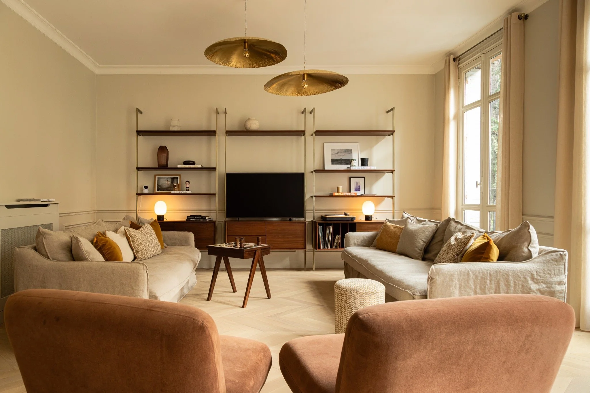 Parisian living room with cream sofas, ochre cushions, terracotta lounge chairs, open wooden shelving and gold-toned pendants.