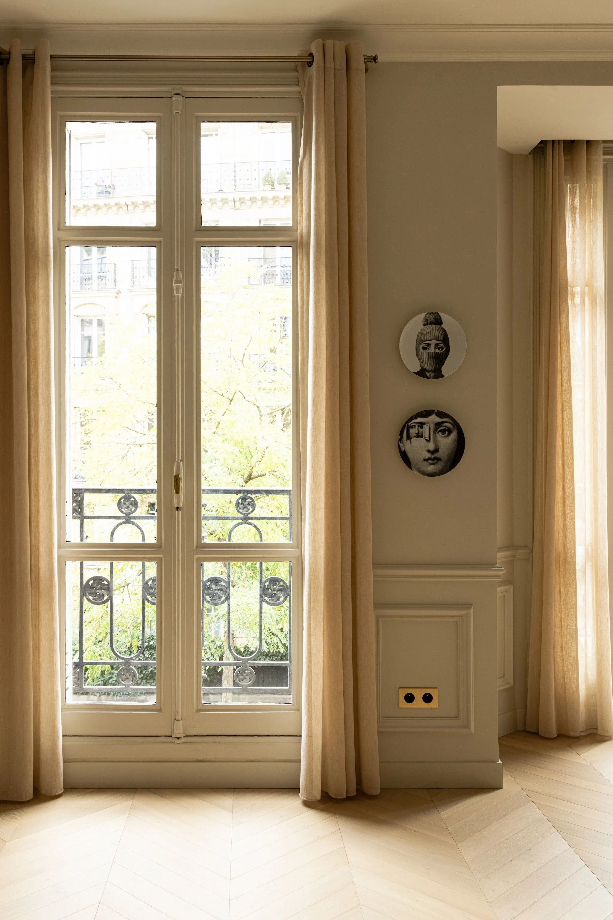 Tall French window with cream curtains, decorative balcony railings and two black-and-white face plates above wall mouldings.