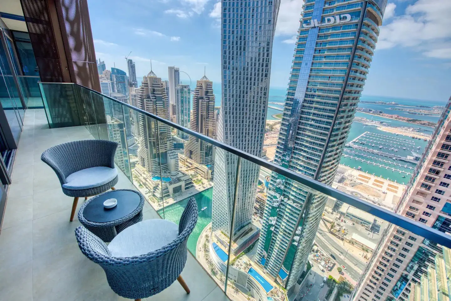 Dubai Marina balcony with two woven armchairs, a round table and glass balustrade overlooking towers and the harbour.