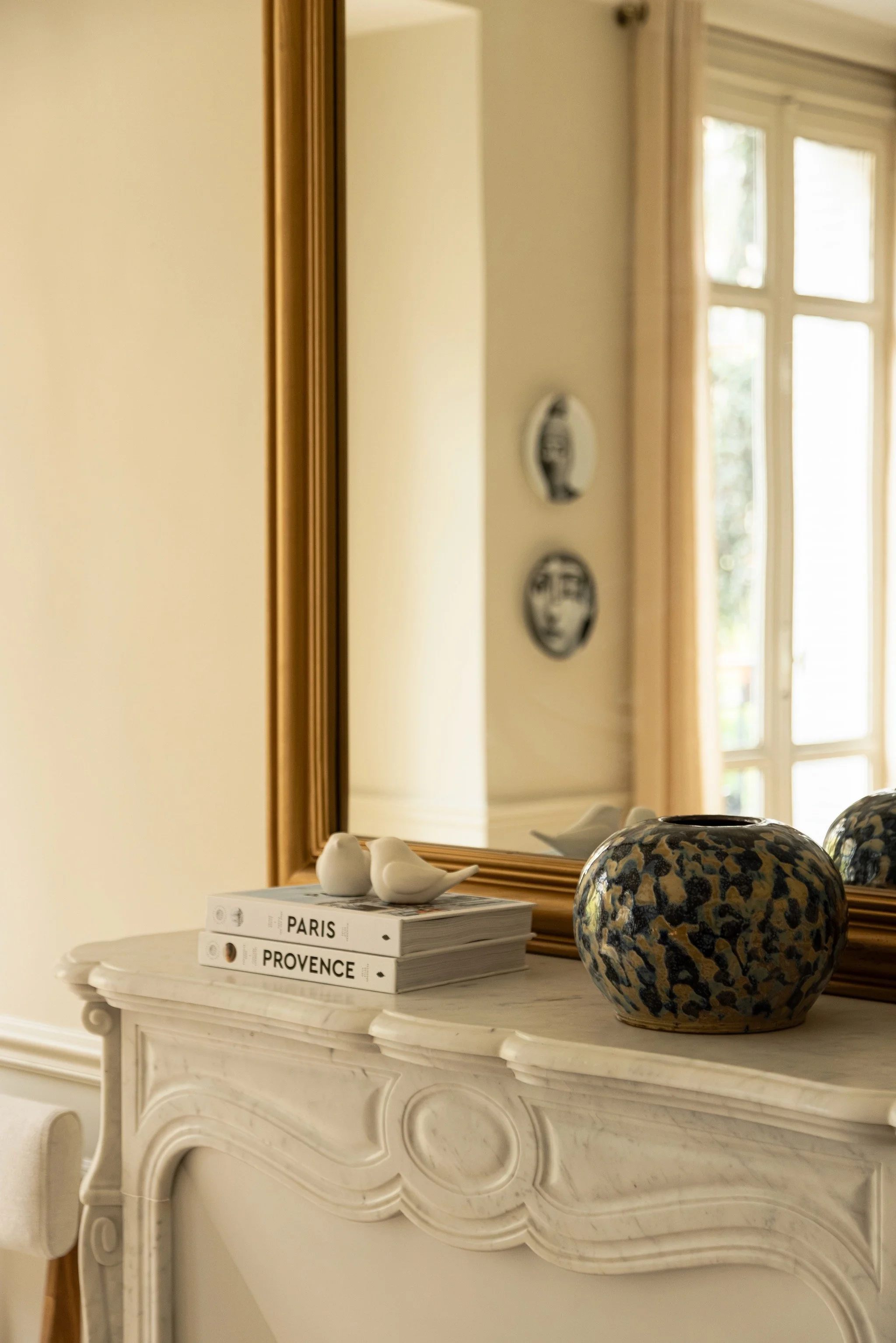 Carved white fireplace mantel with a mottled vase and small bird ornaments beneath a gold-framed mirror.