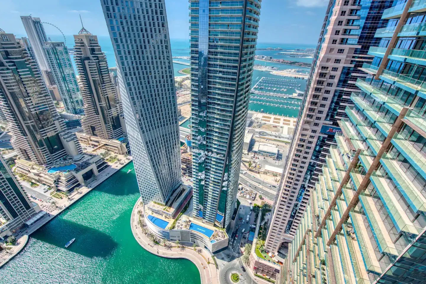 High-angle Dubai Marina view with turquoise water, waterfront towers, roads and harbour berths beyond.