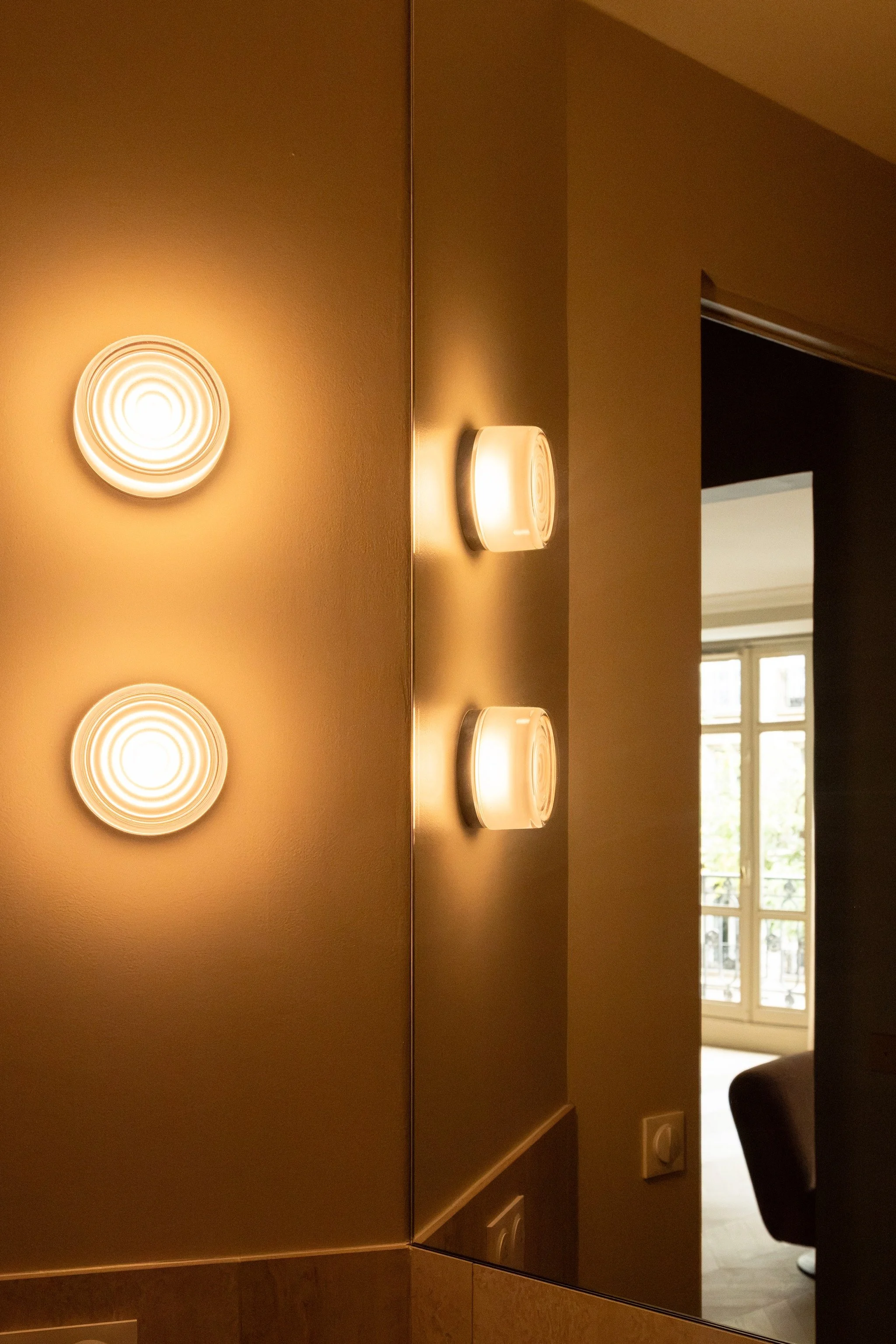 Two glowing circular wall lights reflected in a large mirror, with warm ochre walls and a view towards the living room.