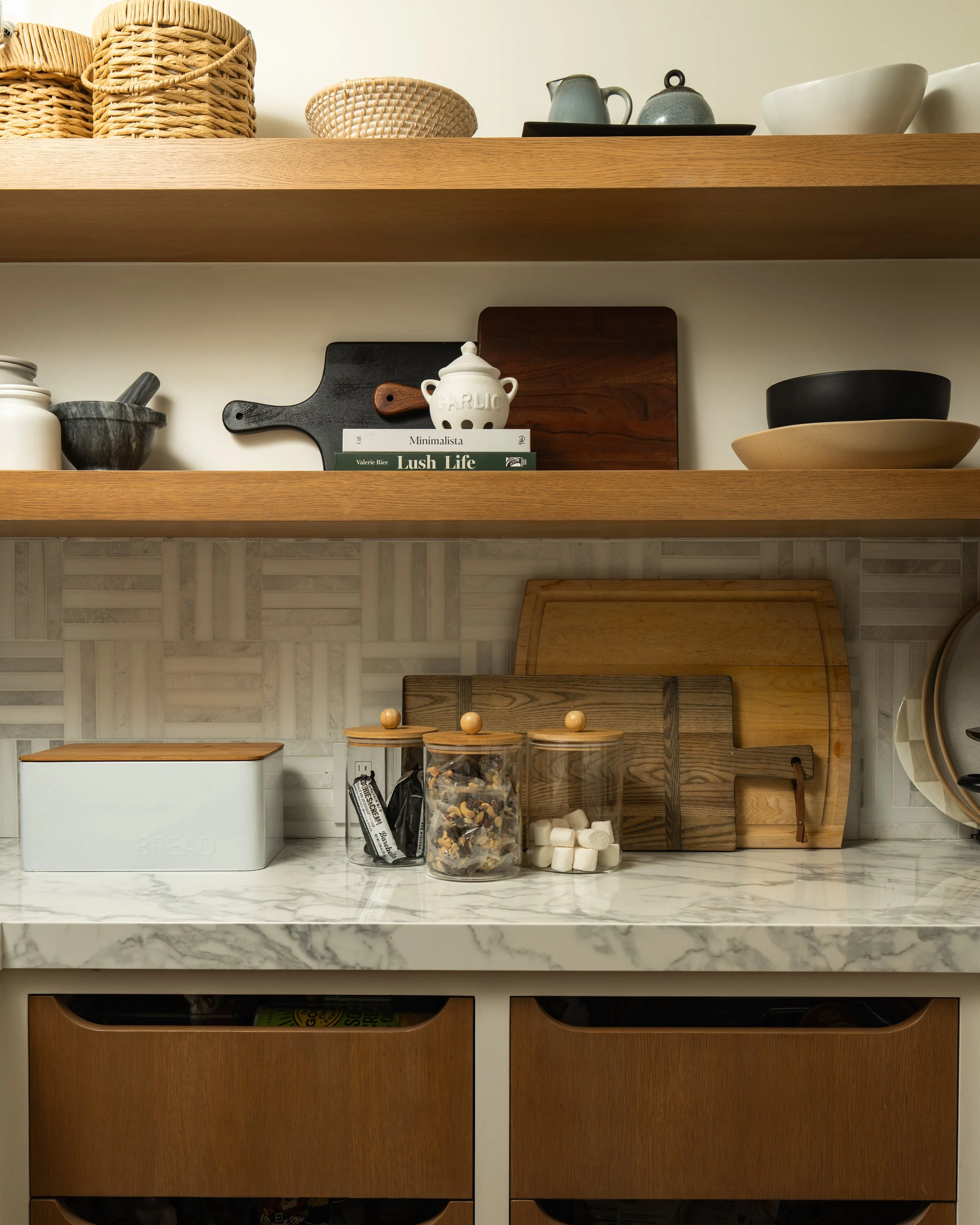 Open kitchen shelves with woven baskets, ceramic bowls, a small cream pitcher, books, cutting boards, mortar and pestle, jars, and containers on marble countertop with a mosaic tile backsplash.