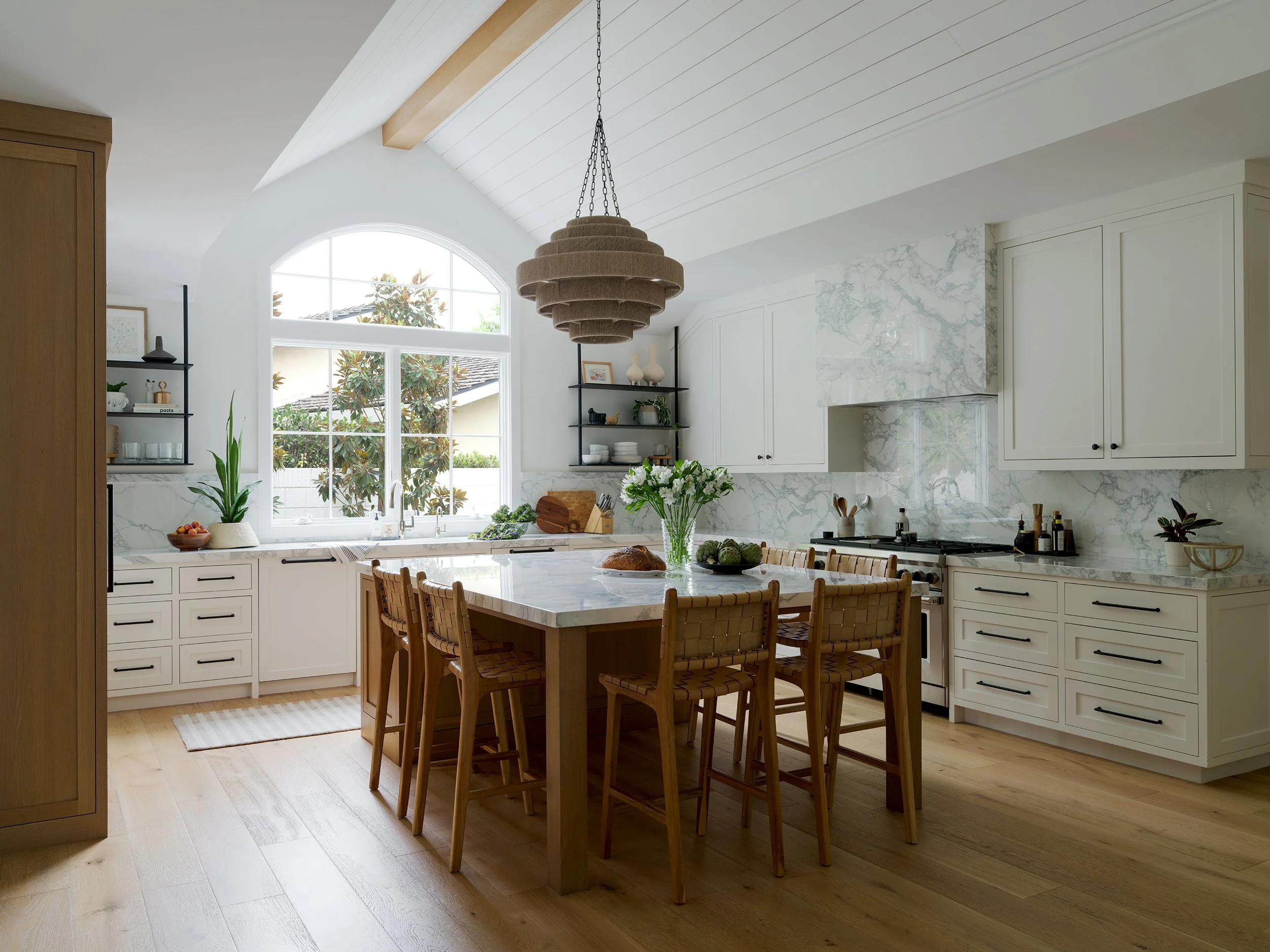 A bright kitchen with white cabinets, marble backsplash, and a kitchen island with a wooden table and chairs. Large arched window with natural sunlight and greenery outside. Overhead, a layered beige pendant light hangs from the ceiling.