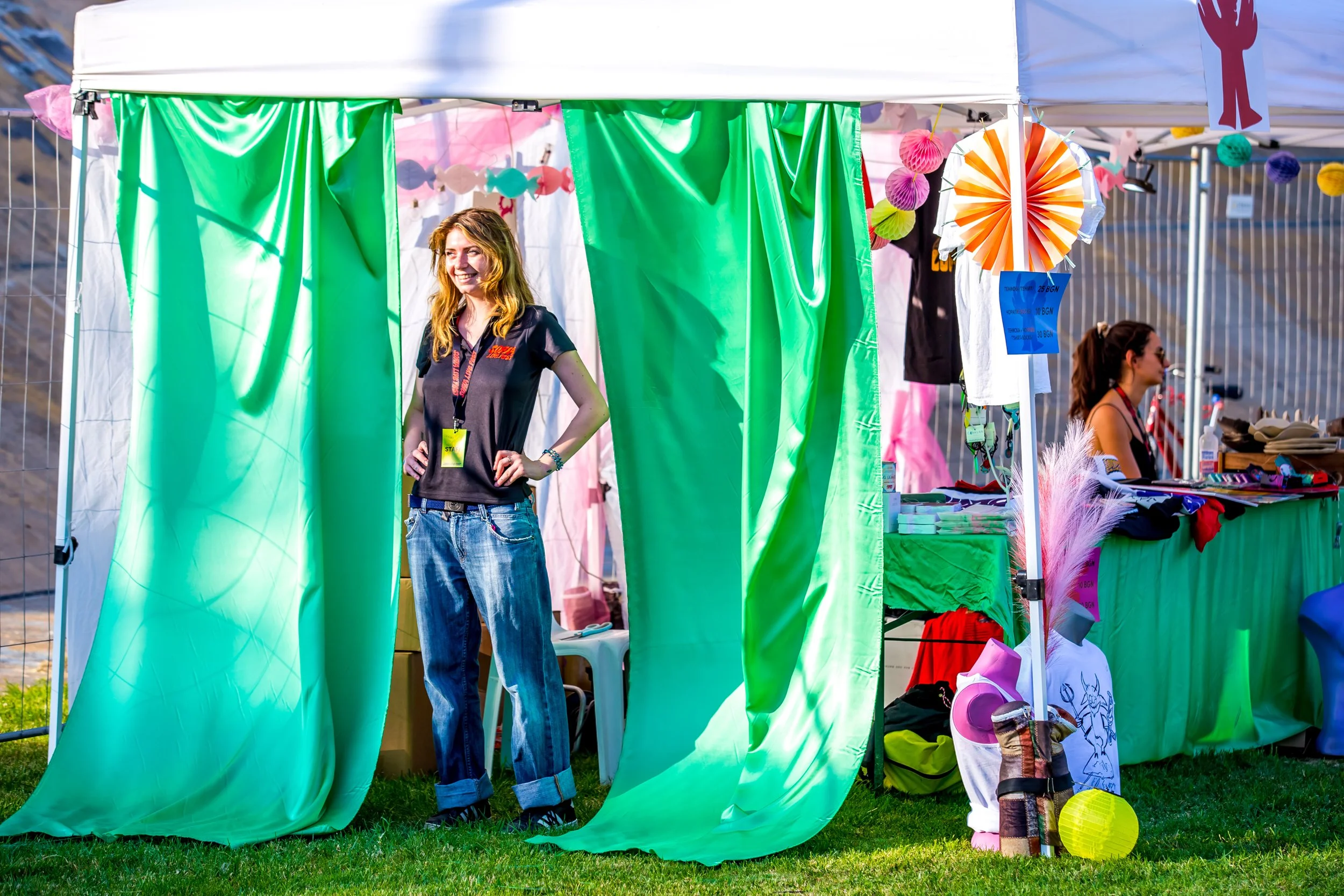Outdoor booth with green curtains, woman standing inside smiling, various decorations and items displayed, another person seated at a table.