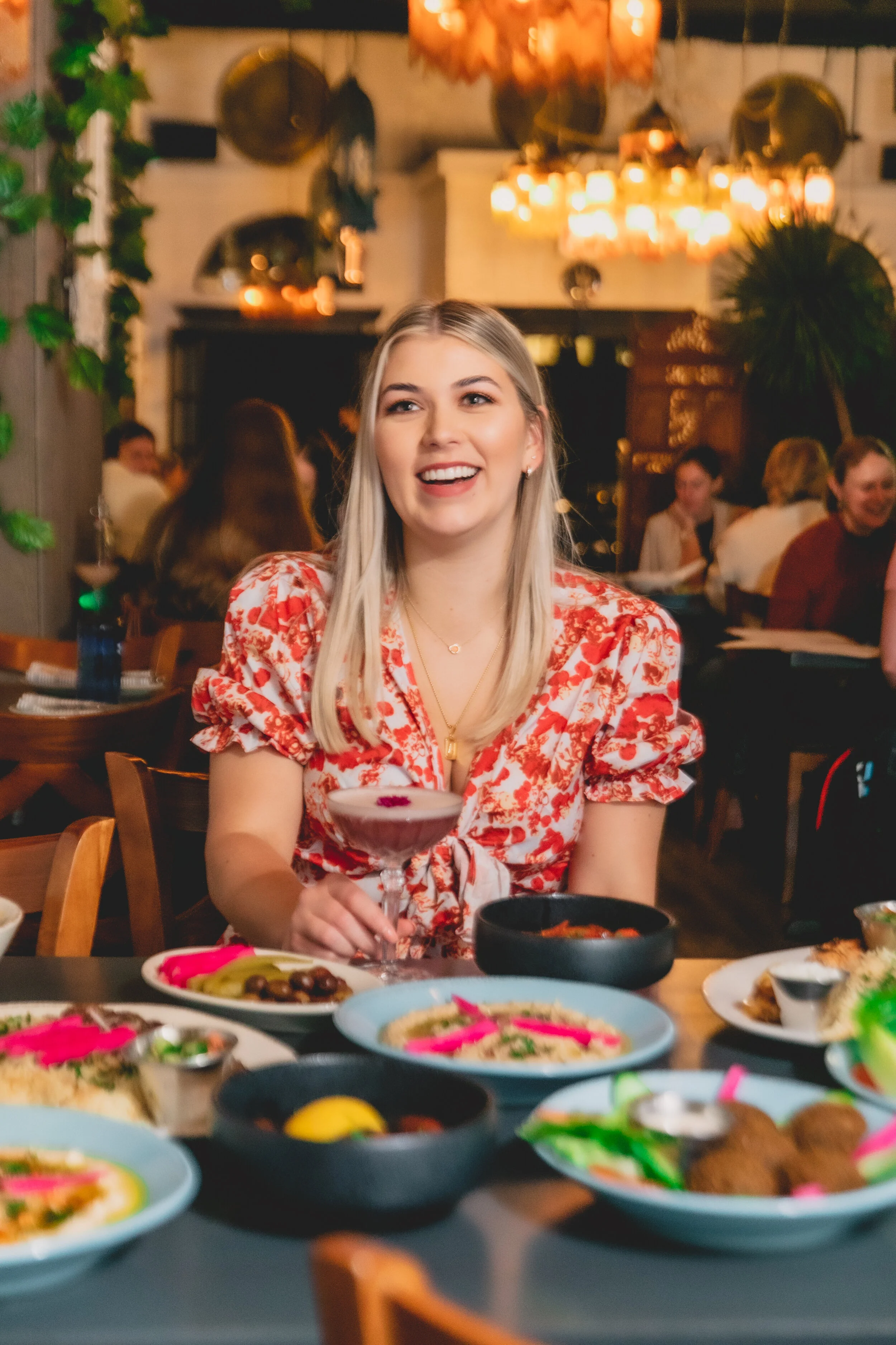 A young woman with blonde hair sitting at a table in a restaurant, smiling and holding a cocktail. The table is filled with various dishes, including bowls of food and appetizers, with warm lighting overhead.
