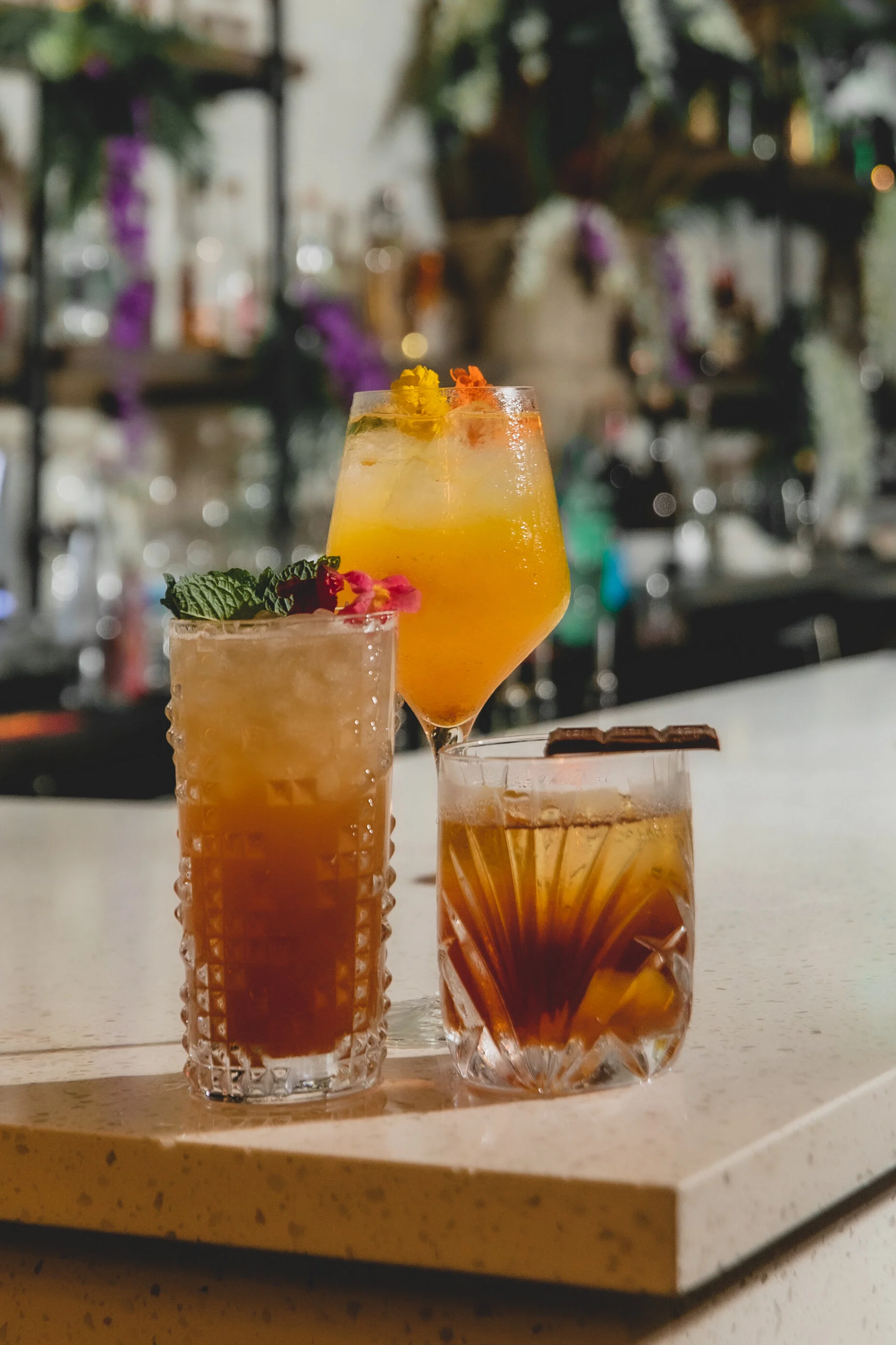 Three cocktails on a white counter with a blurred bar background, featuring colorful drinks garnished with herbs and fruits.