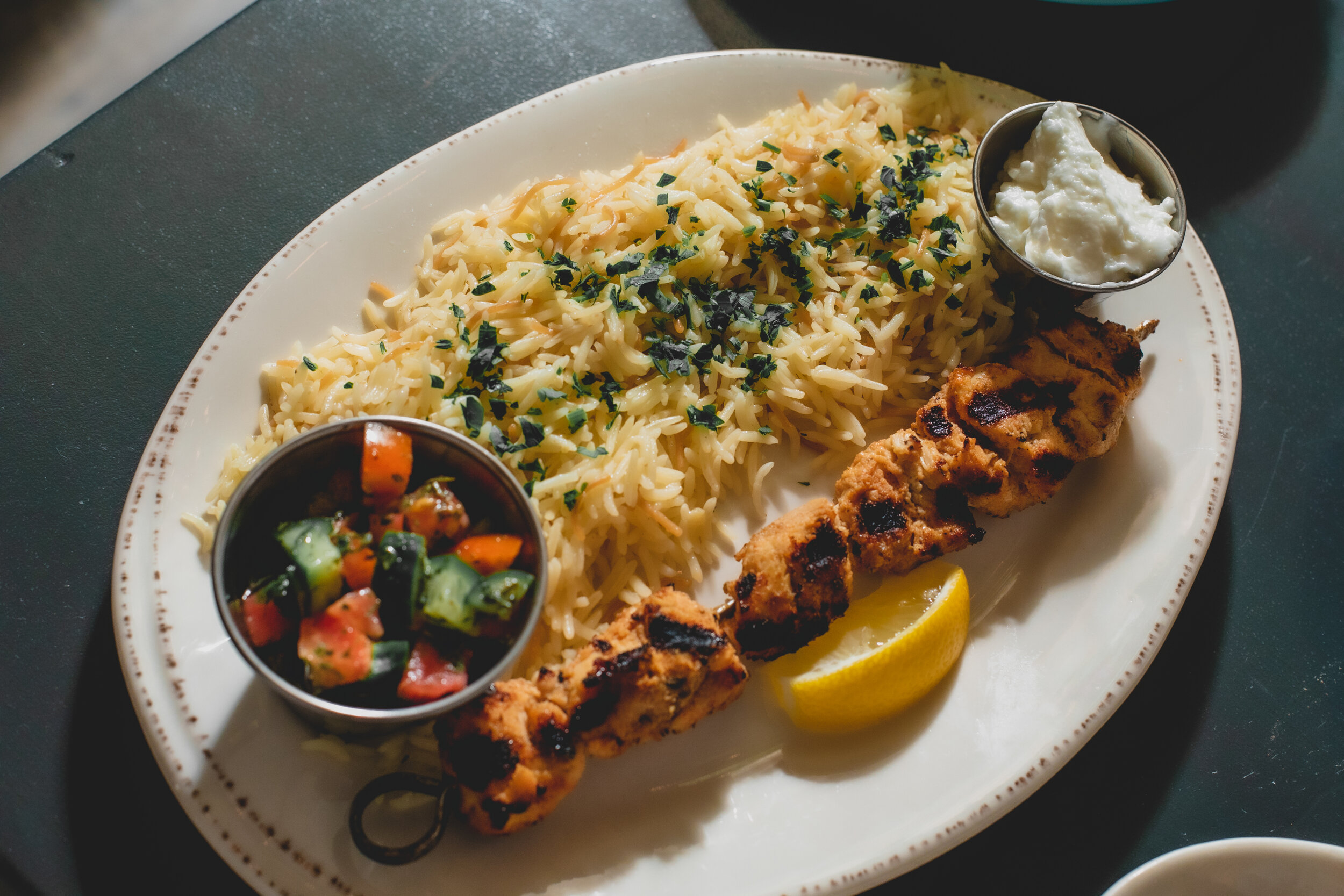 Plate of Indian cuisine with grilled chicken skewer, basmati rice topped with herbs, cucumber and tomato salad, mint chutney, and raita, garnished with a lemon wedge.