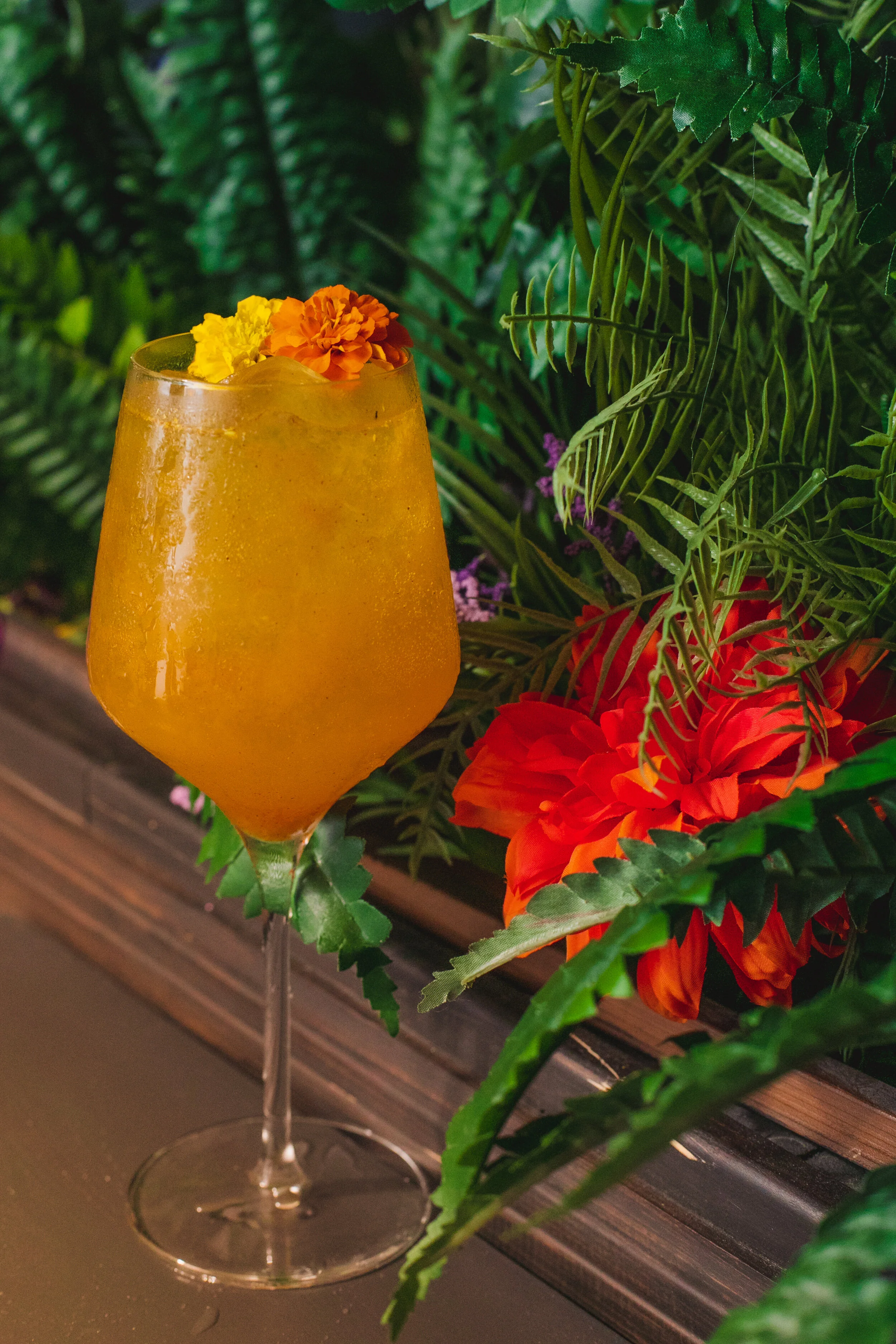 A tall glass of orange cocktail with ice, garnished with yellow and orange flowers, surrounded by green foliage and red flowers.