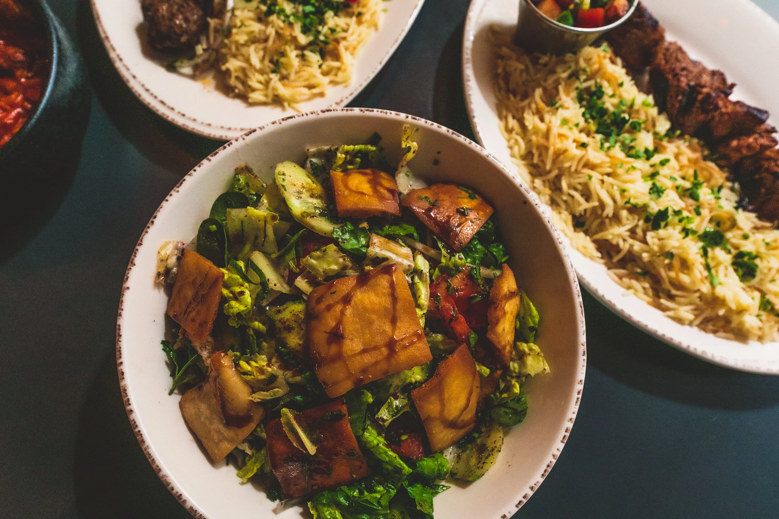 A bowl of salad with chopped cucumbers, tomatoes, and grilled eggplant chunks, with bowls of rice and grilled meat on plates in the background.