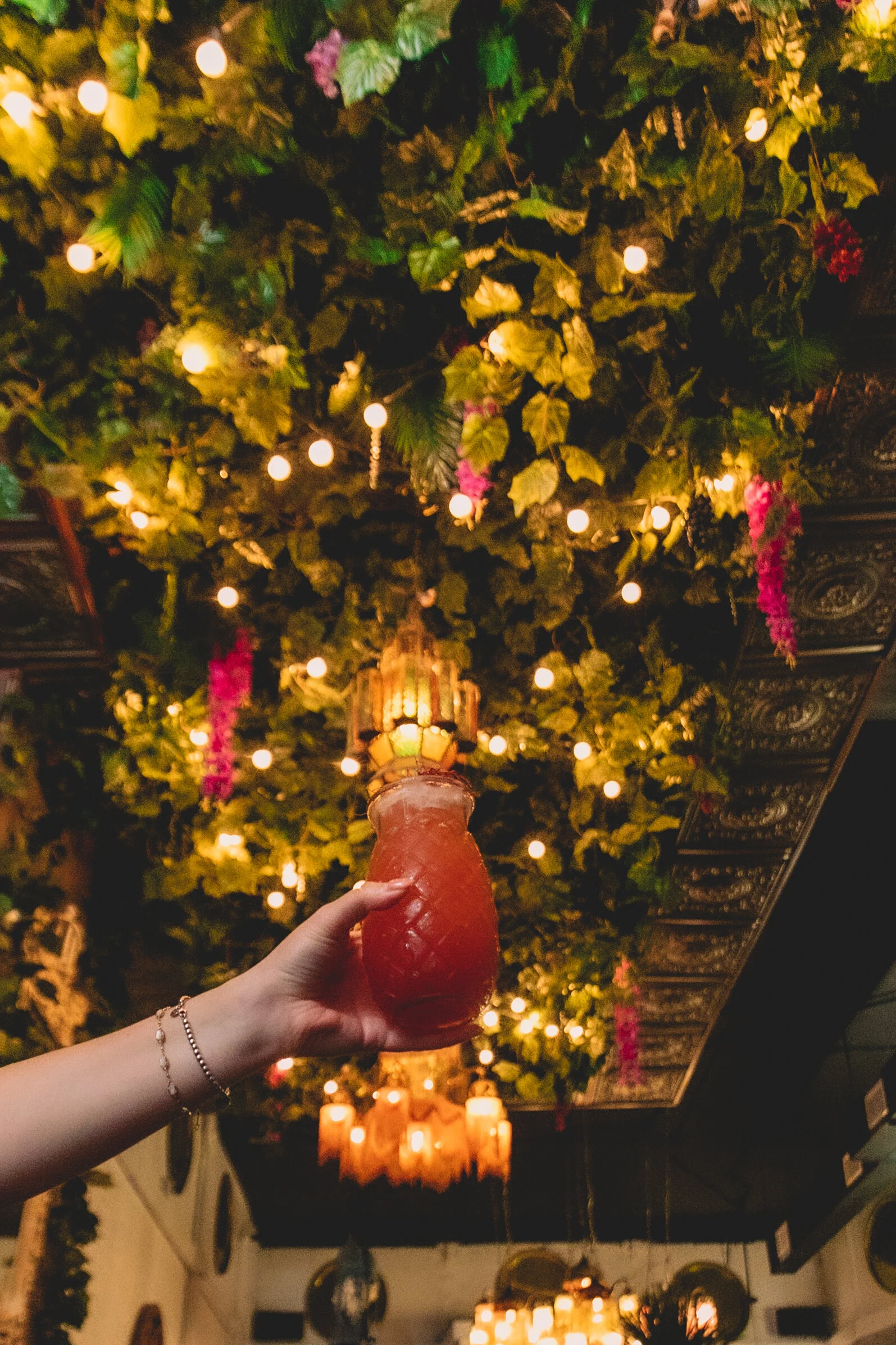 A person holding a red, pineapple-shaped drink with a straw, in a dimly lit restaurant decorated with hanging lights, lush greenery, and colorful flowers on the ceiling.