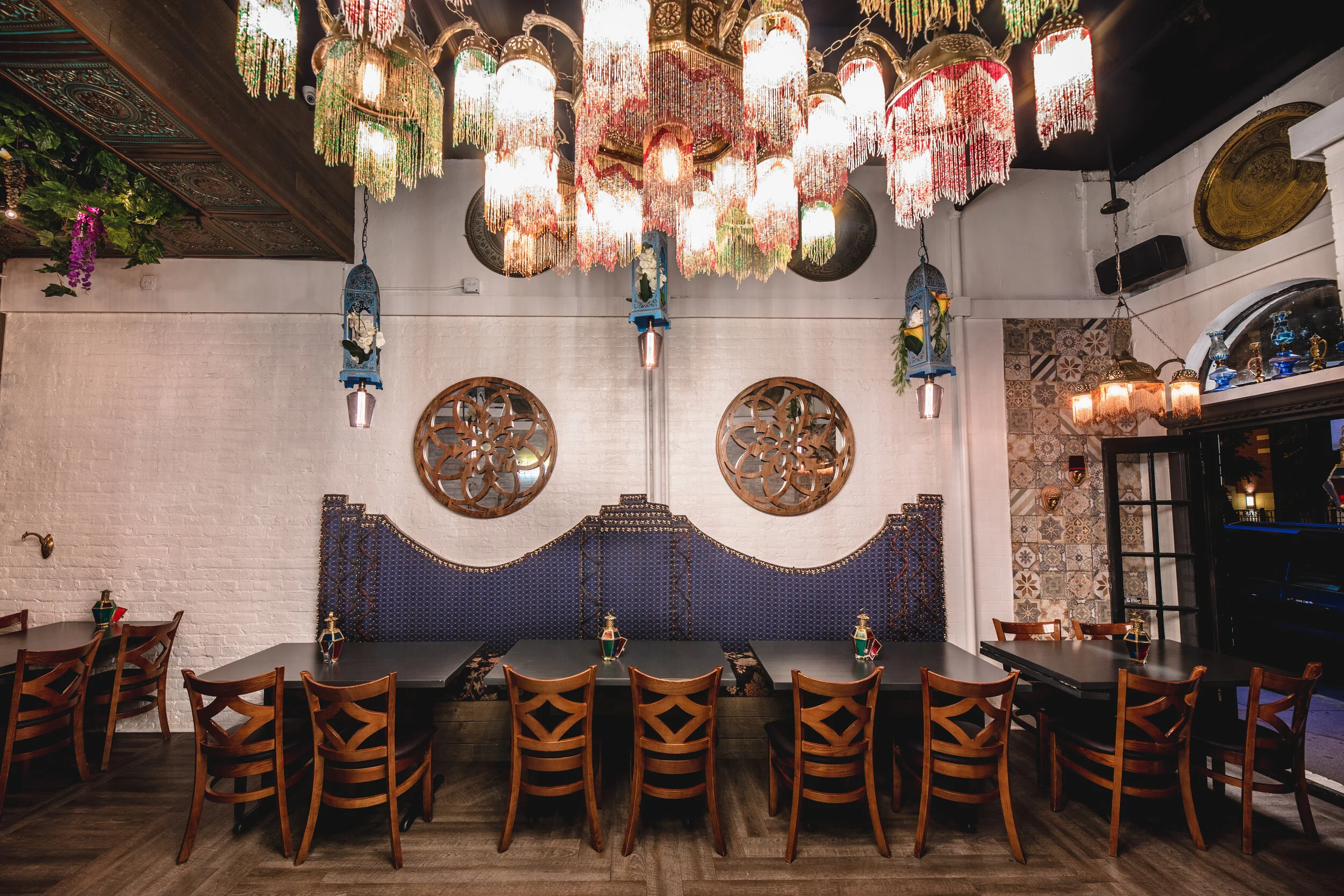 Interior of a restaurant with a white brick wall, decorated with round wooden art pieces and patterned tiles. There are multiple tables with wooden chairs, some with small bottles on top. The ceiling features colorful, tasseled light fixtures, and tr