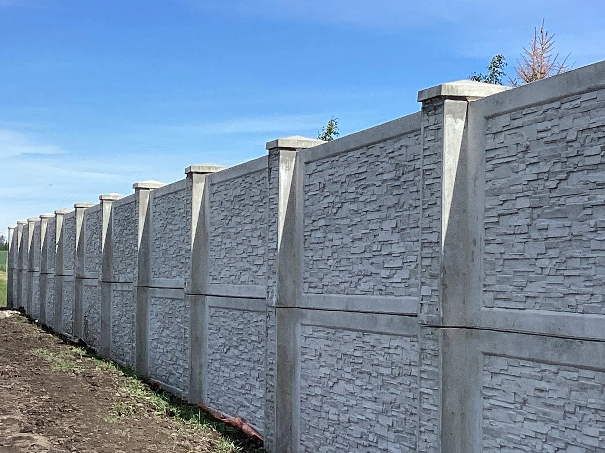 A long concrete wall with textured stone pattern panels and concrete caps on top, set against a blue sky with a few trees in the background.