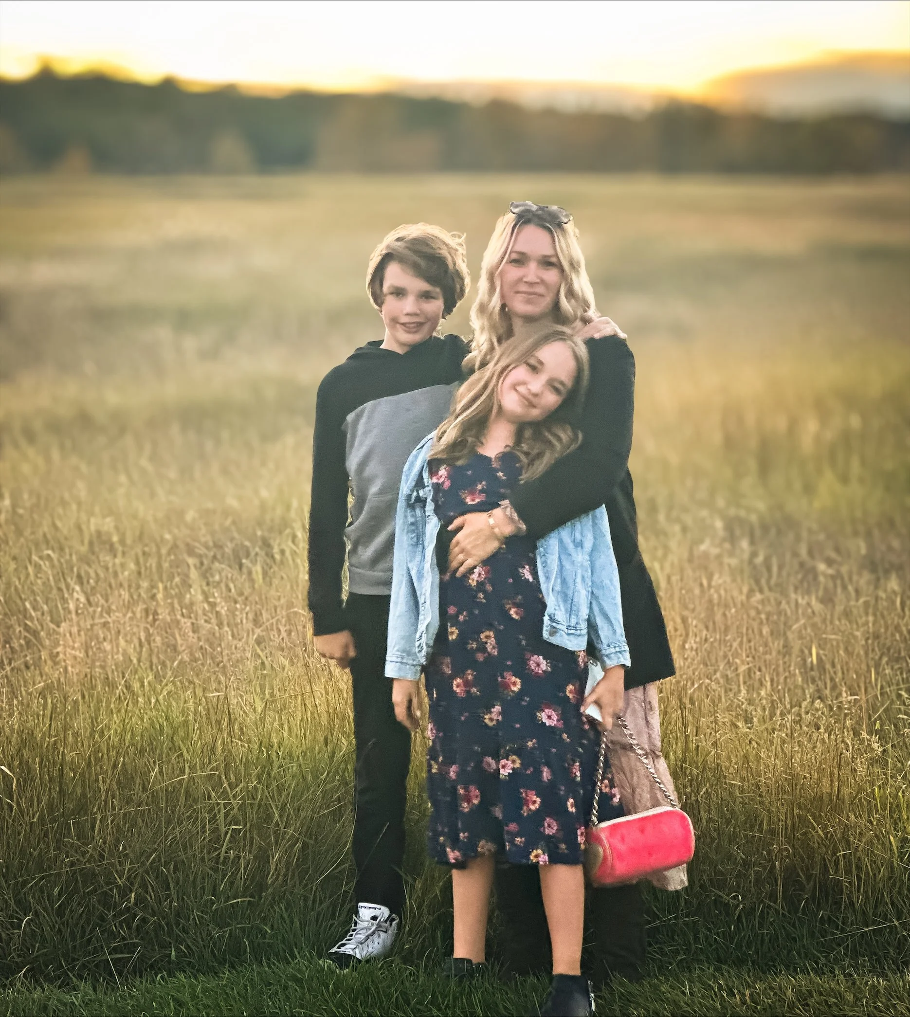 A woman with two children standing in a grassy field during sunset, smiling at the camera.