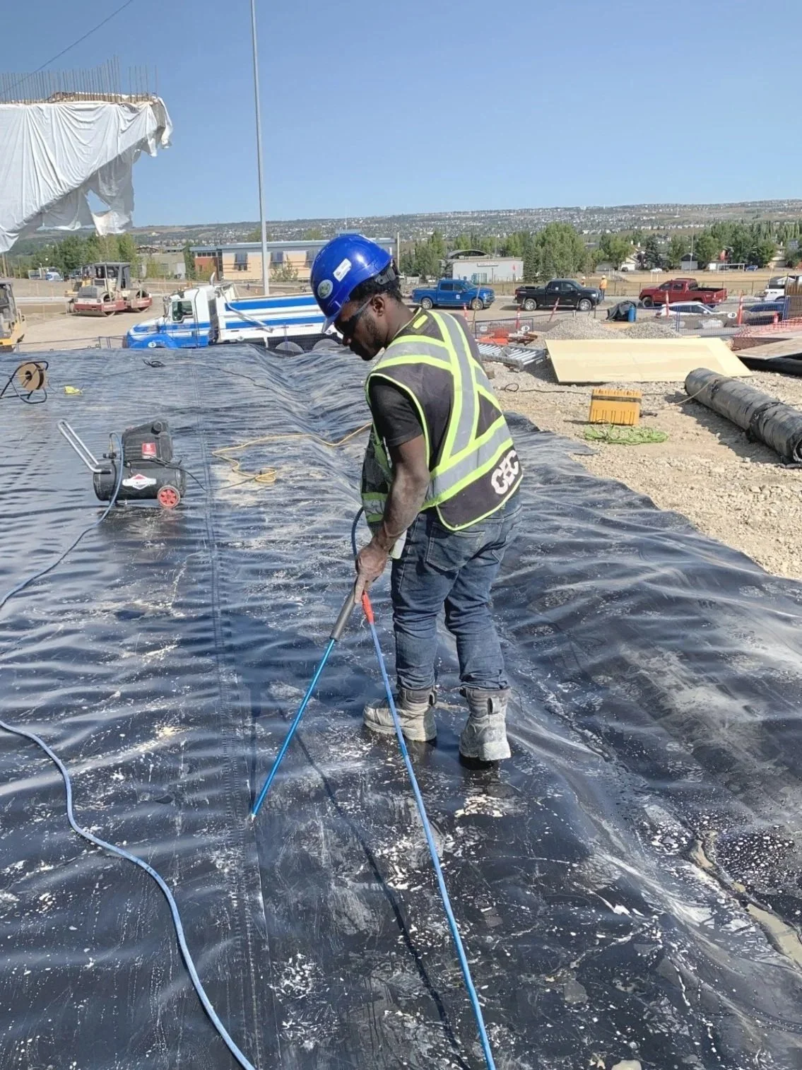 A construction worker wearing a blue safety helmet, sunglasses, and a reflective safety vest is working on a black waterproof membrane on a building roof. He is using a tool to cut or secure the membrane, with construction equipment and vehicles in the background.