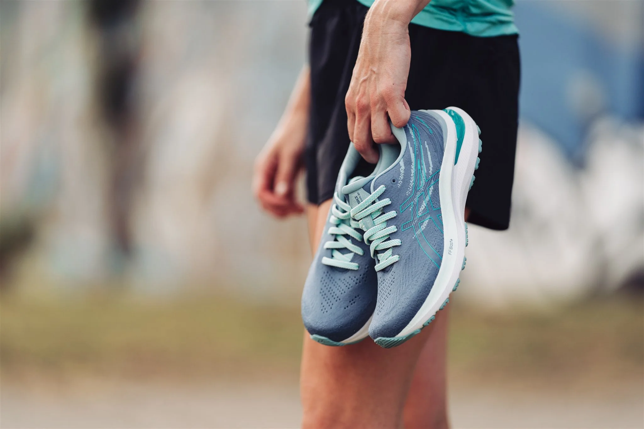 Person standing outdoors, holding a pair of blue running shoes in their hand.