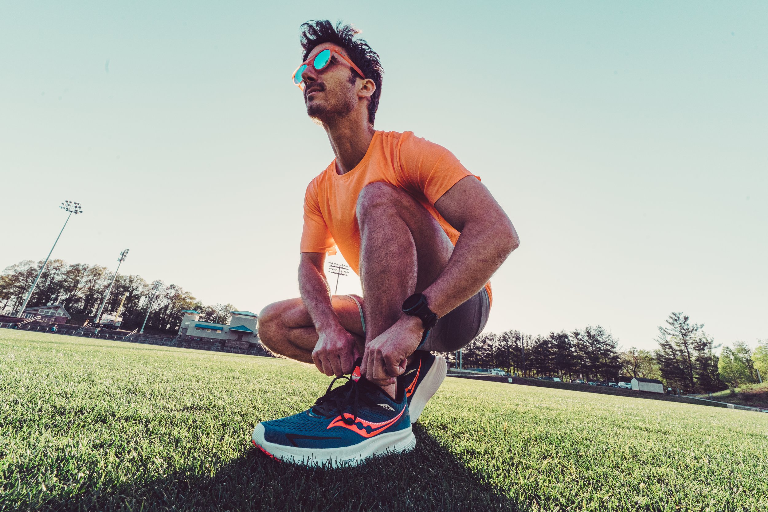 A man in an orange shirt and sunglasses is tying his running shoe on a grassy track field at an outdoor stadium during daylight.
