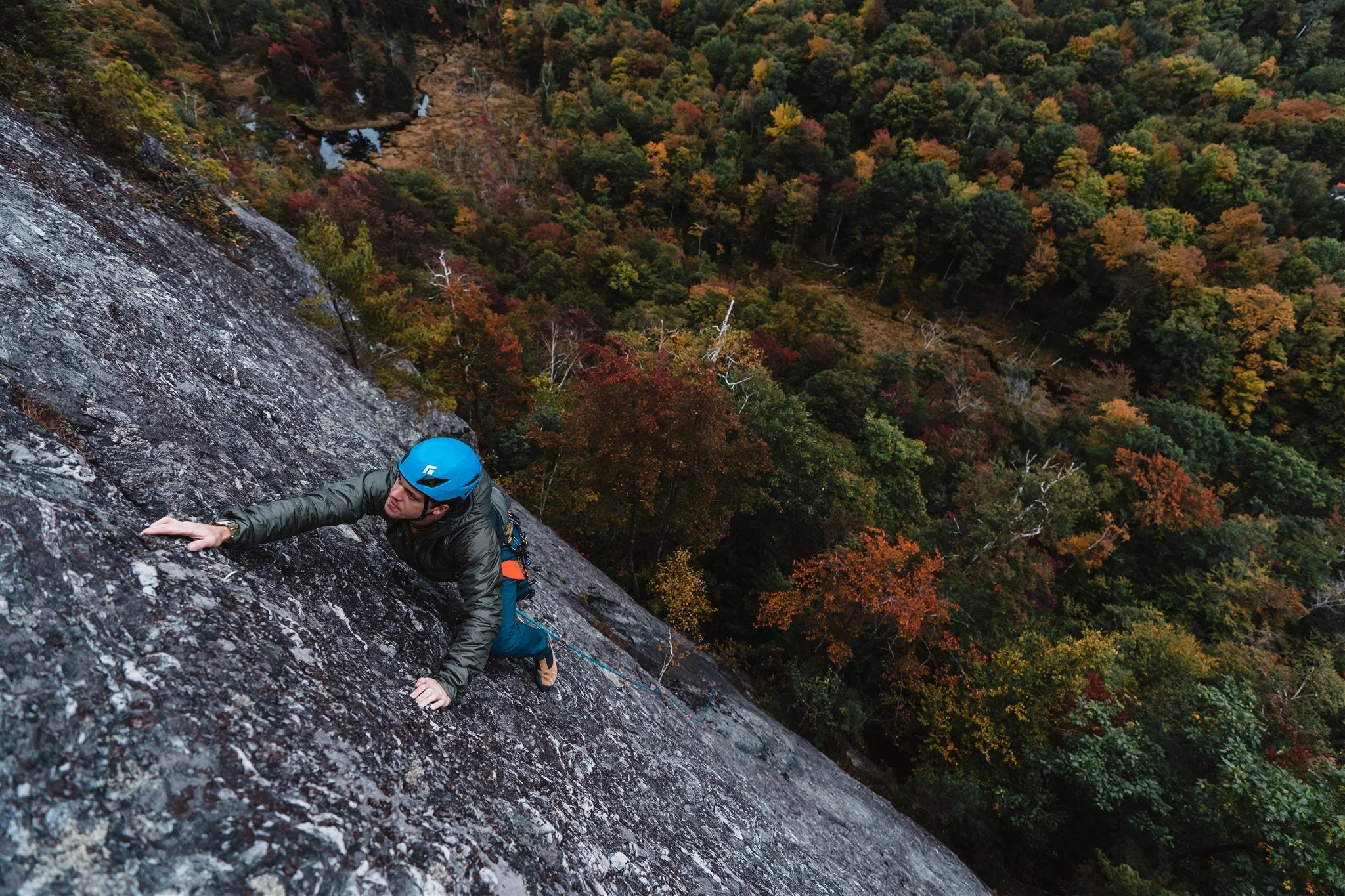 A male rock climber wearing a blue helmet and gray jacket ascending a steep rock face with a forested landscape below