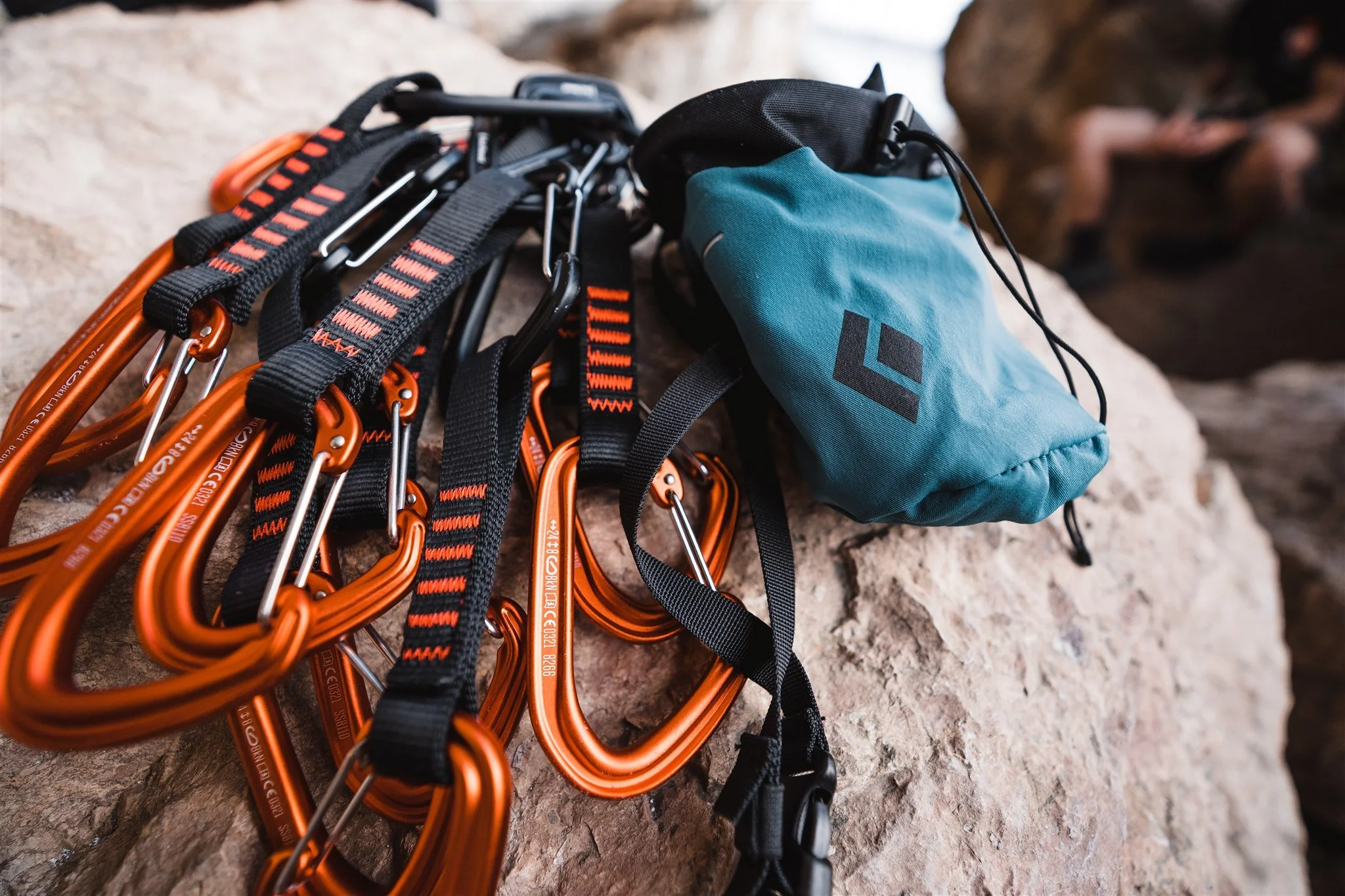Climbing gear including orange carabiners and straps with a blue chalk bag on a rocky surface, with a person in the background sitting on a rock.