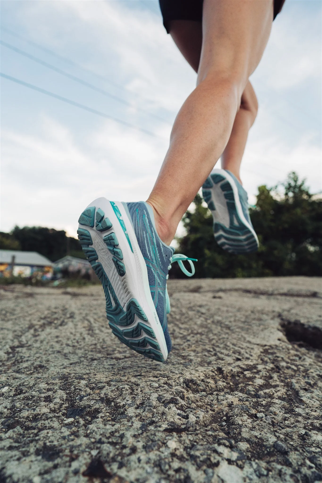 Close-up of a person's legs and feet in running shoes, mid-stride on a rocky surface, with trees and a cloudy sky in the background.