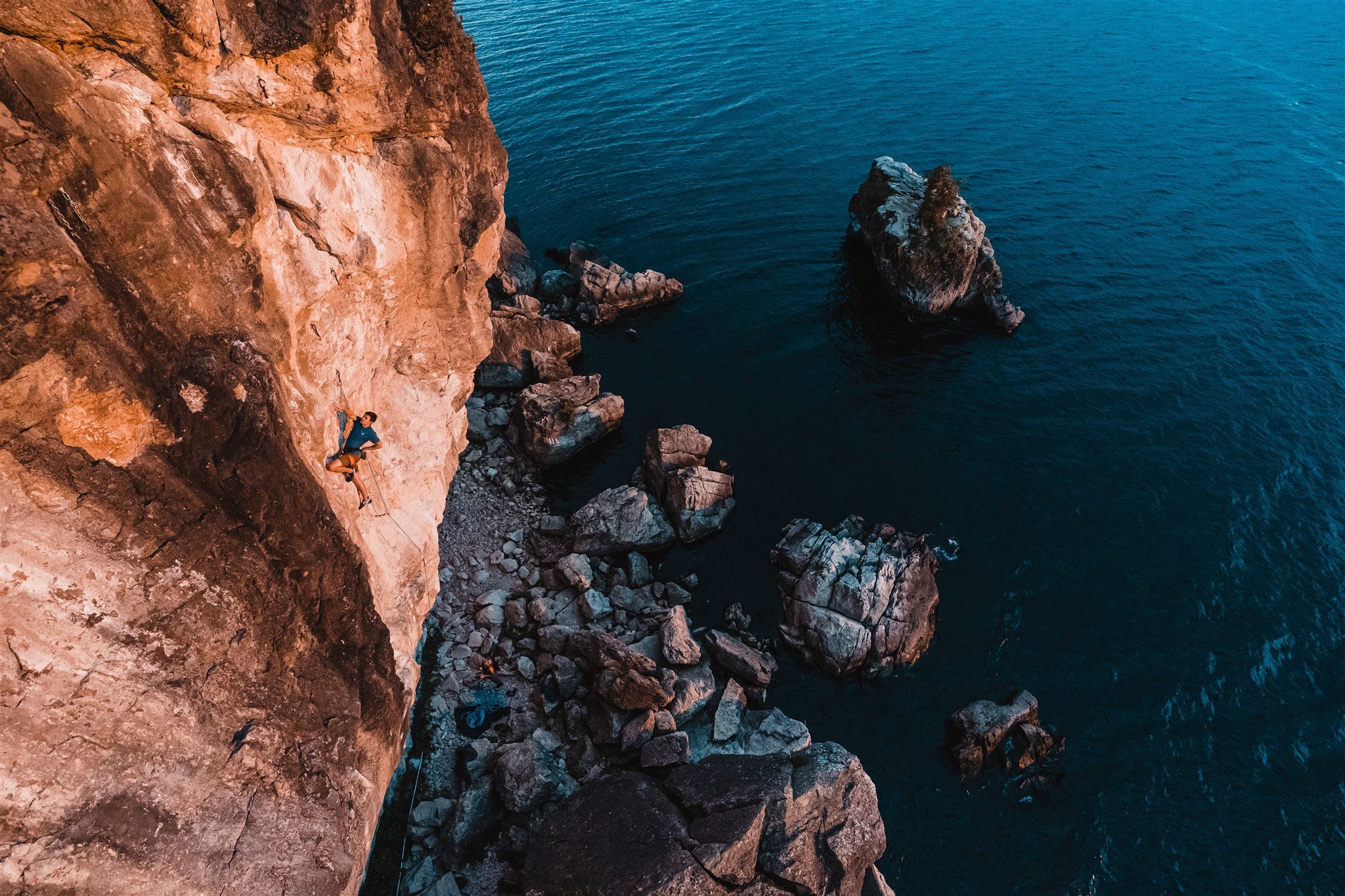 A person rock climbing on a steep cliff face above blue water with large rocks below in the water, during sunset.