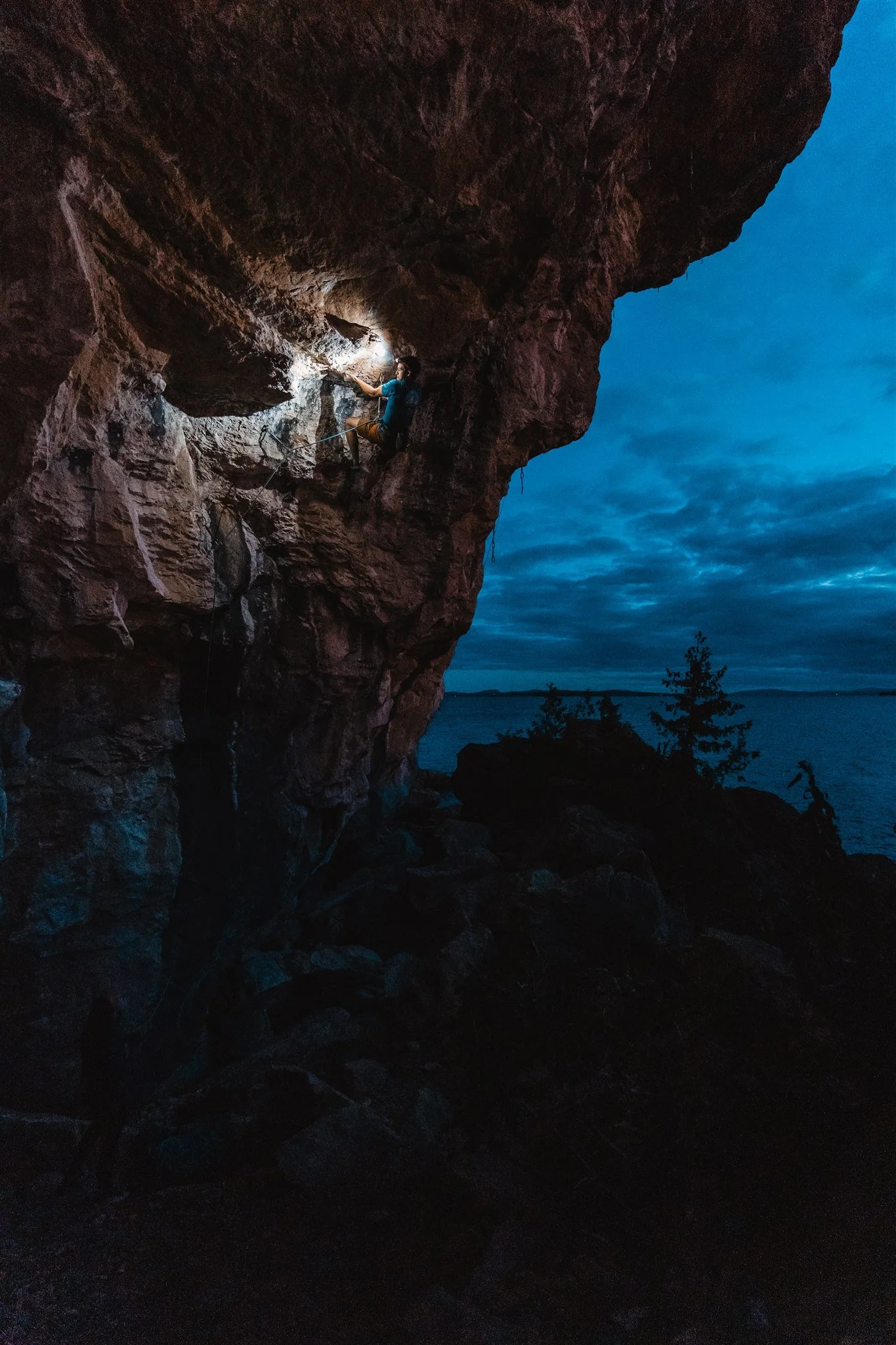 A person camping and climbing a rock face at dusk near a lake, illuminated by a headlamp.