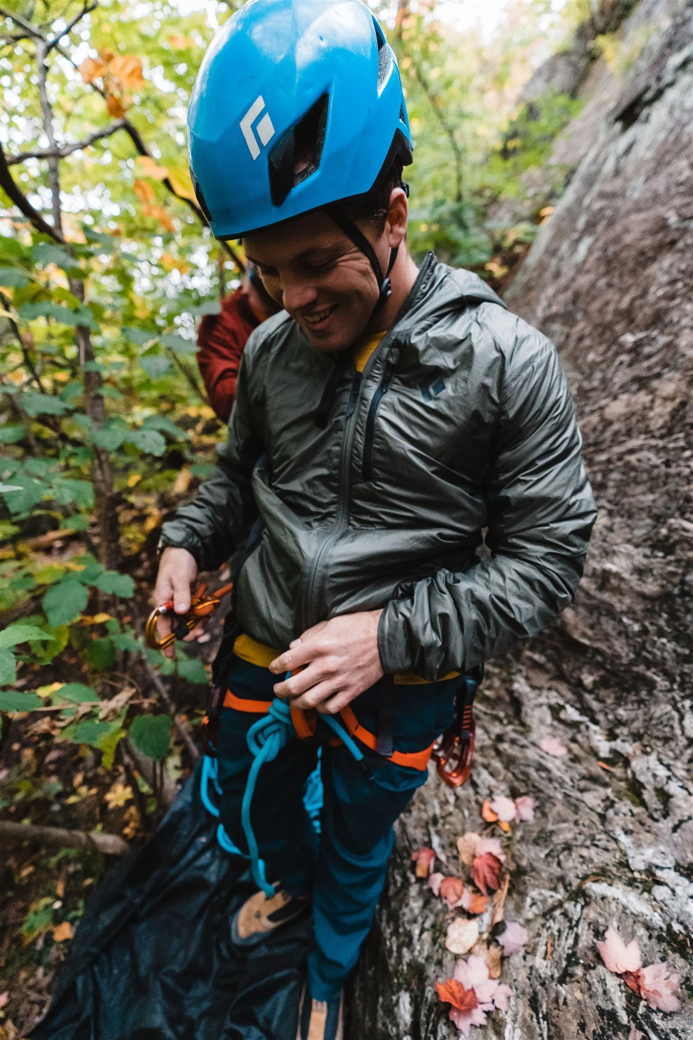 A man in outdoor climbing gear, including a blue helmet and a gray jacket, standing on a rocky ledge in a forest.